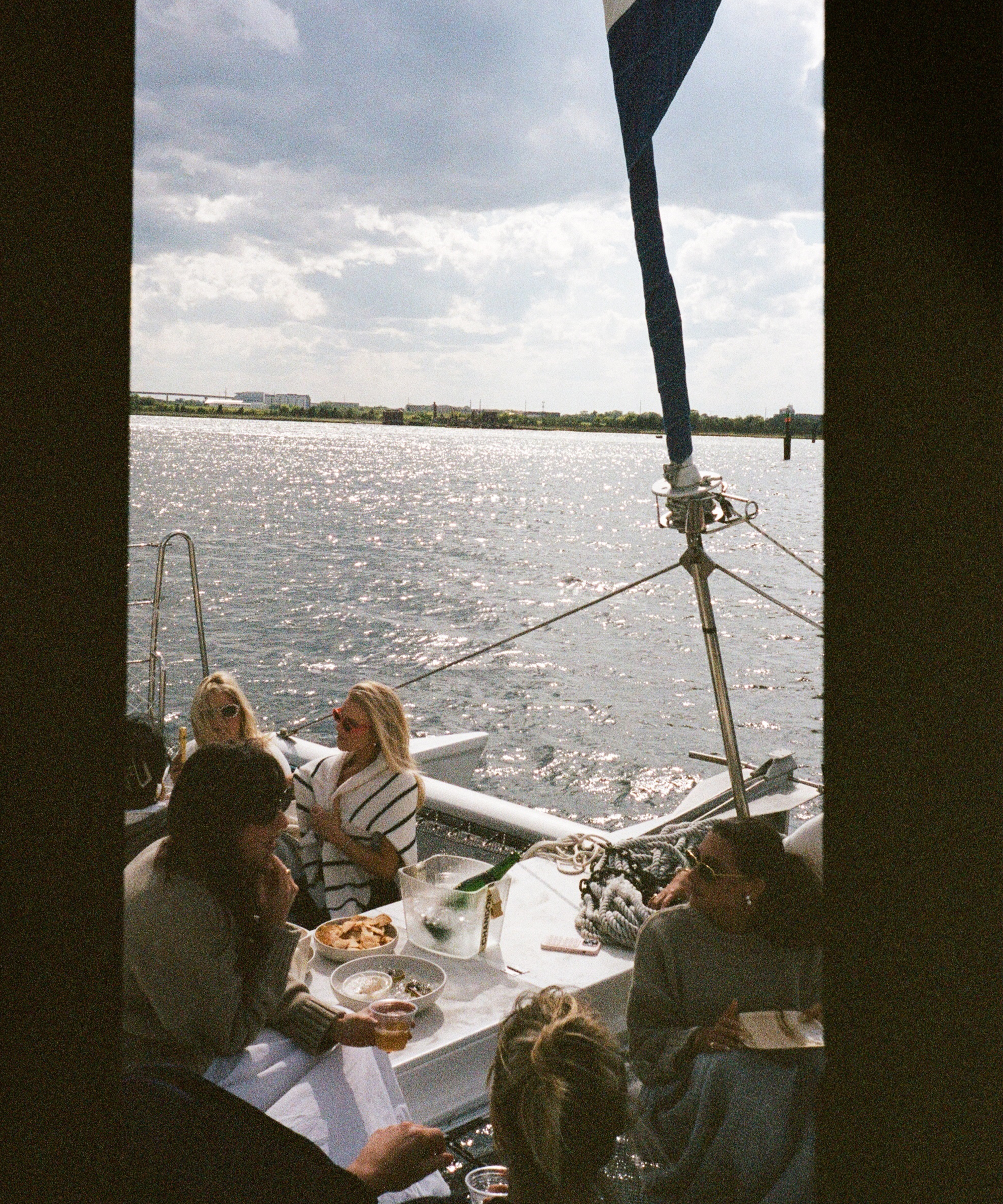 A group of people are sitting and eating on a sailboat near Charleston, enjoying food and drinks on a sunny day with sparkling water in the background. A large flag and boat ropes are visible near the bow.