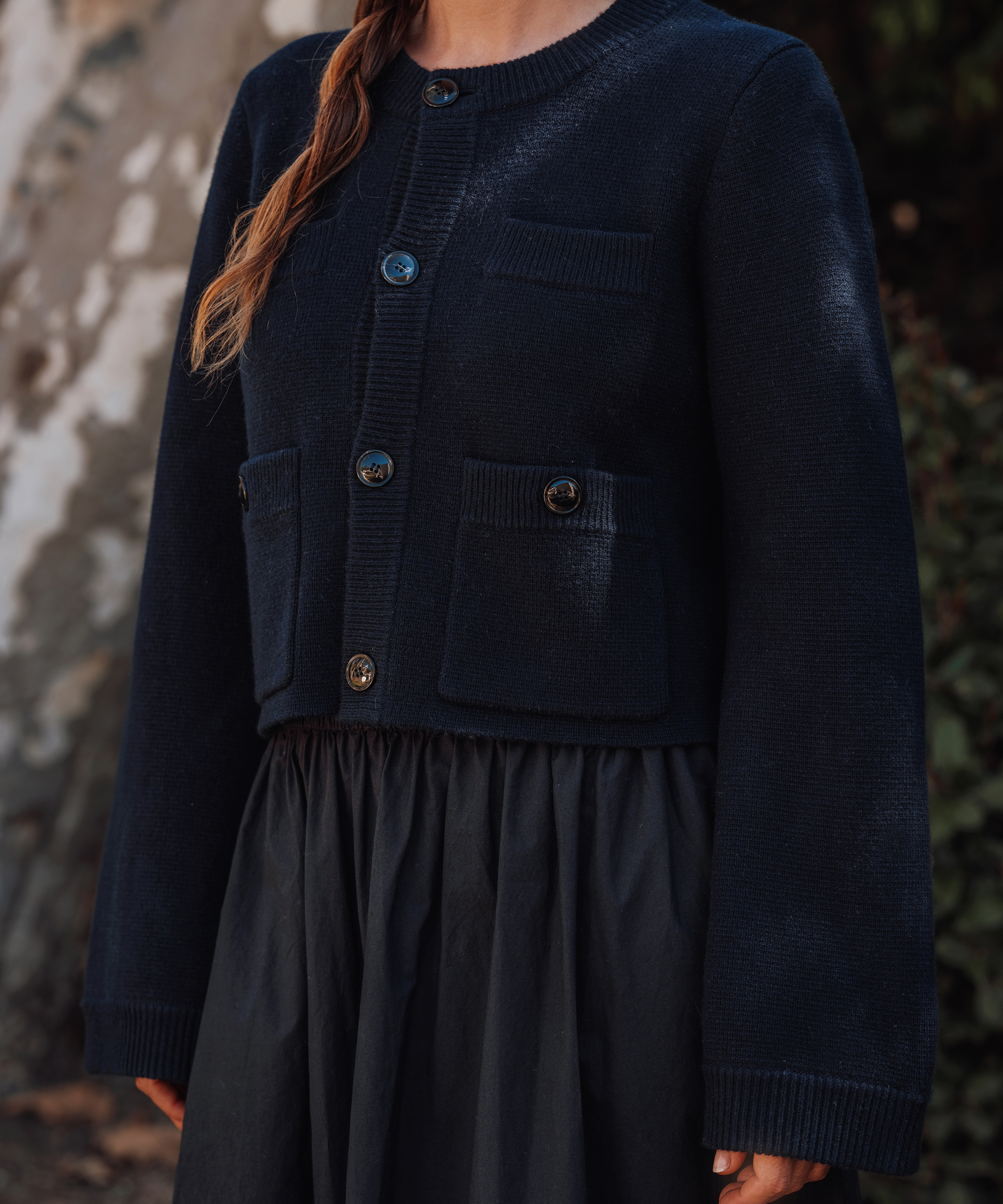 A person wearing a dark navy blue buttoned cardigan with front pockets and a matching skirt, styled as a chic spring to summer outfit, stands outdoors. Their face is not visible in the image.