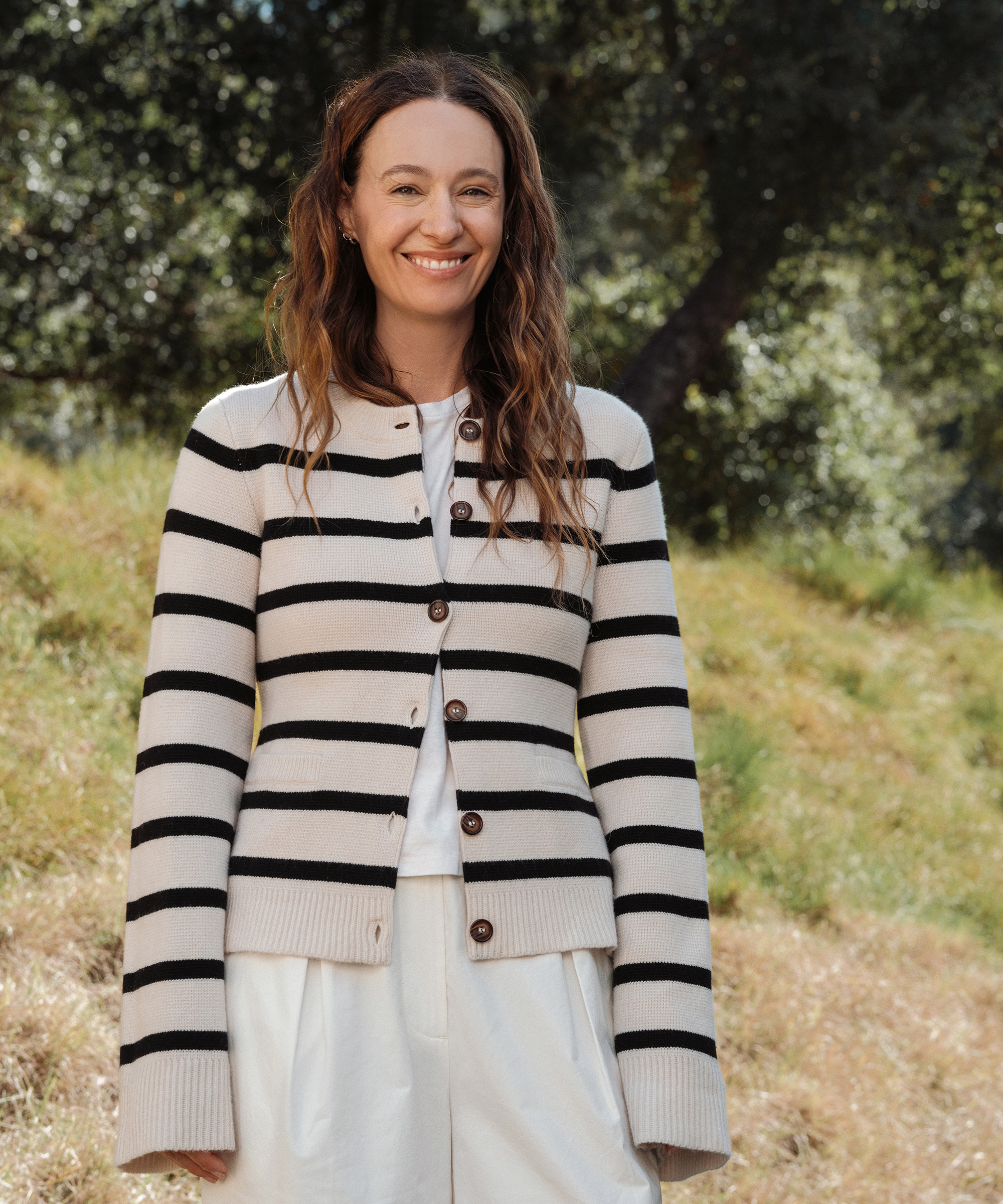 A woman with long wavy hair stands outdoors, smiling in her effortless spring style—a cream sweater with black stripes over a white shirt and white pants, set against sunlit grass and leafy trees.