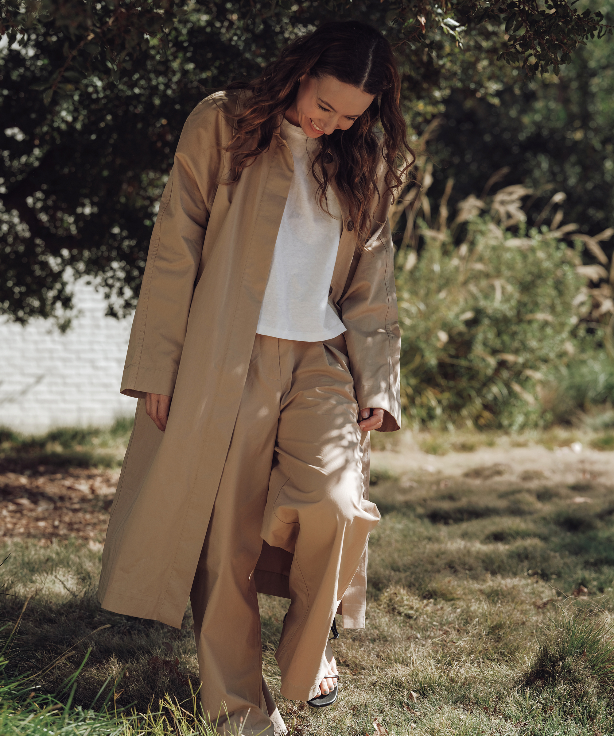 A woman stands outdoors on grass, looking down and smiling. Showcasing effortless spring style, she wears a long beige coat, matching wide-leg pants, a white shirt, and sandals. Sunlight filters through the trees behind her as her loose wavy hair catches the light.