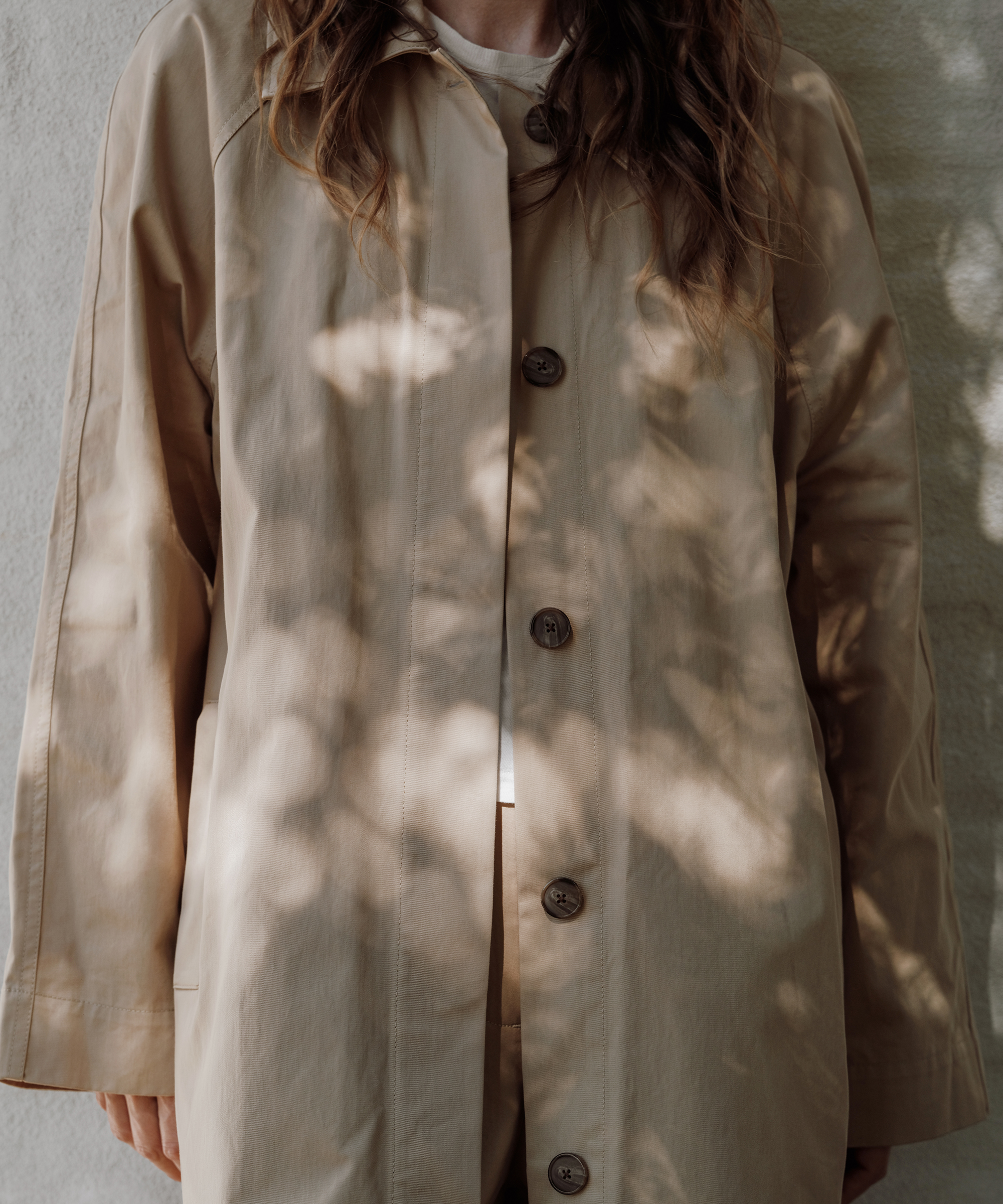A person wearing a beige button-up coat stands against a light wall, with dappled sunlight and shadows creating patterns across the fabric—a subtle nod to effortless spring style. Their face is not visible in the image.