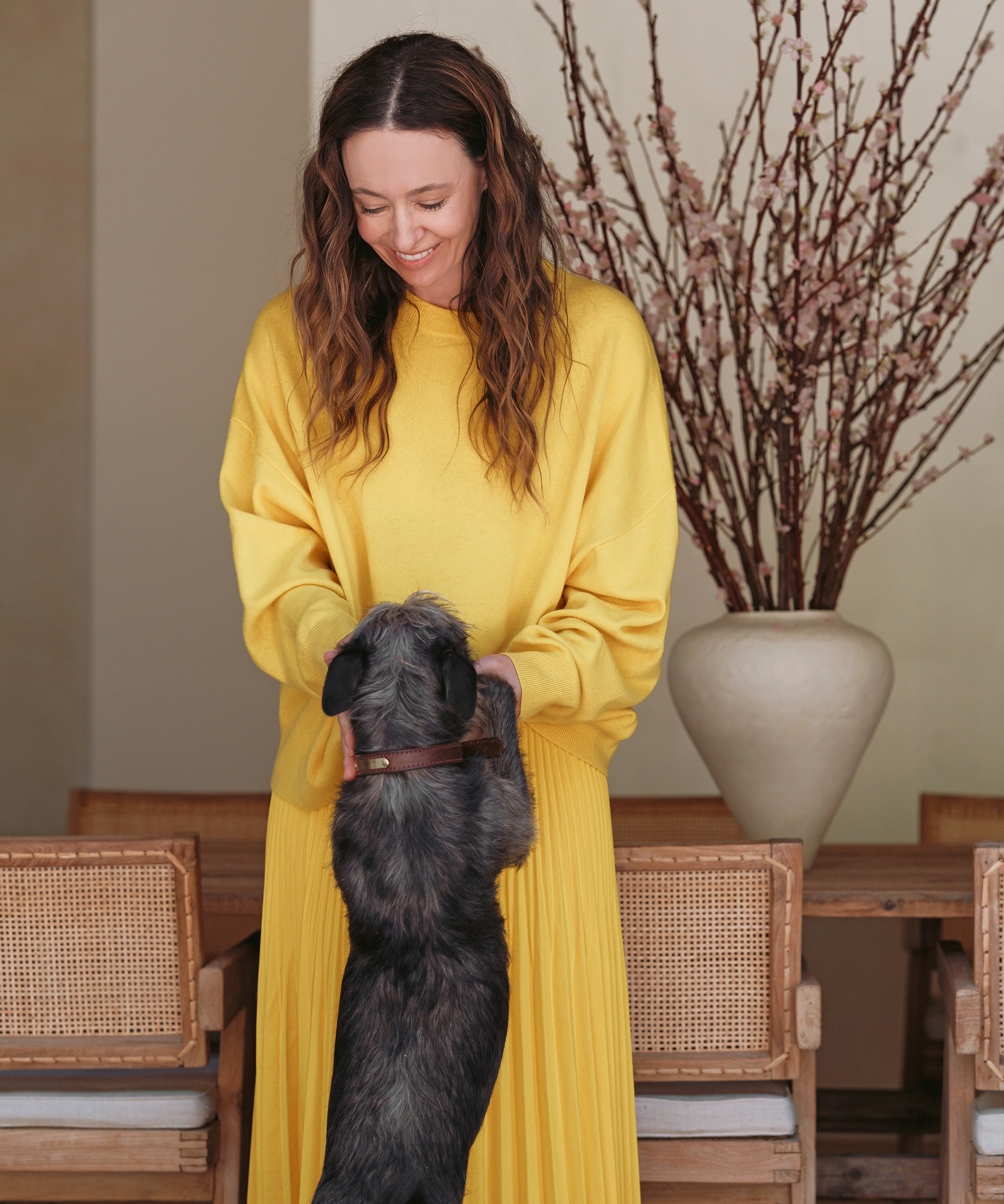 A smiling woman in a bright yellow outfit showcases her spring style indoors, holding a small black dog standing on its hind legs. Behind her, a wooden dining table, wicker chairs, and a large vase with pink branches complete the scene.