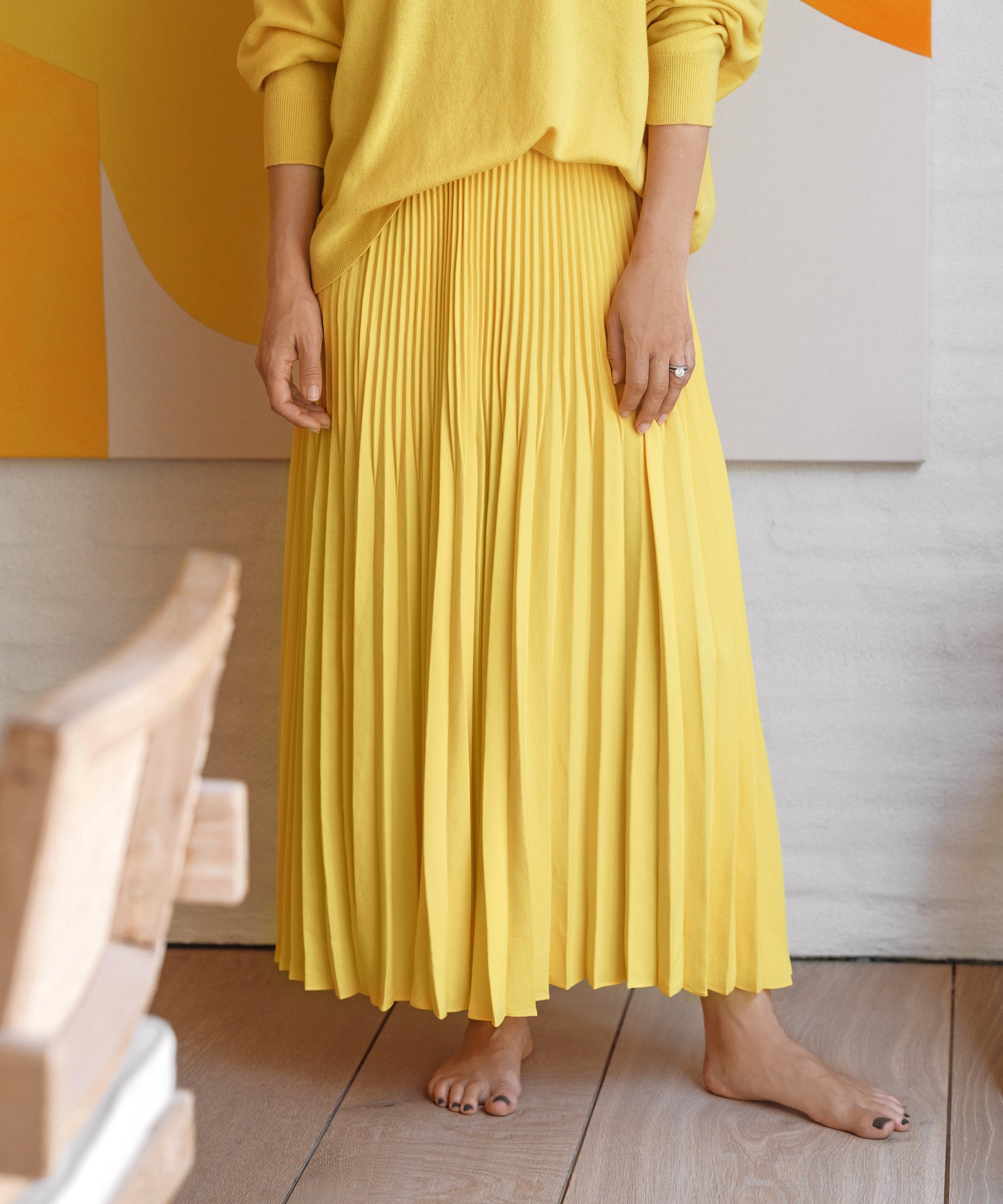 A barefoot person showcases effortless spring style in a bright yellow pleated skirt and matching sweater, standing indoors on a wooden floor. Their hands rest at their sides, framed by the backdrop of a partial abstract artwork.
