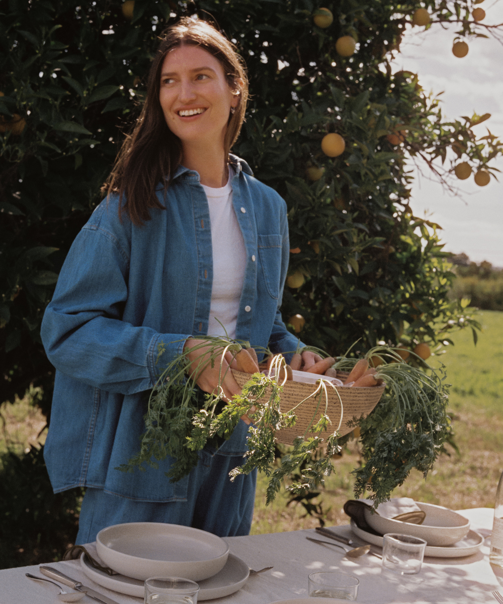 Michèle Ouellet Benson stands outdoors by an orange tree, smiling in her blue denim outfit and holding a basket of fresh carrots. A table set with plates, glasses, and cutlery is arranged before her.