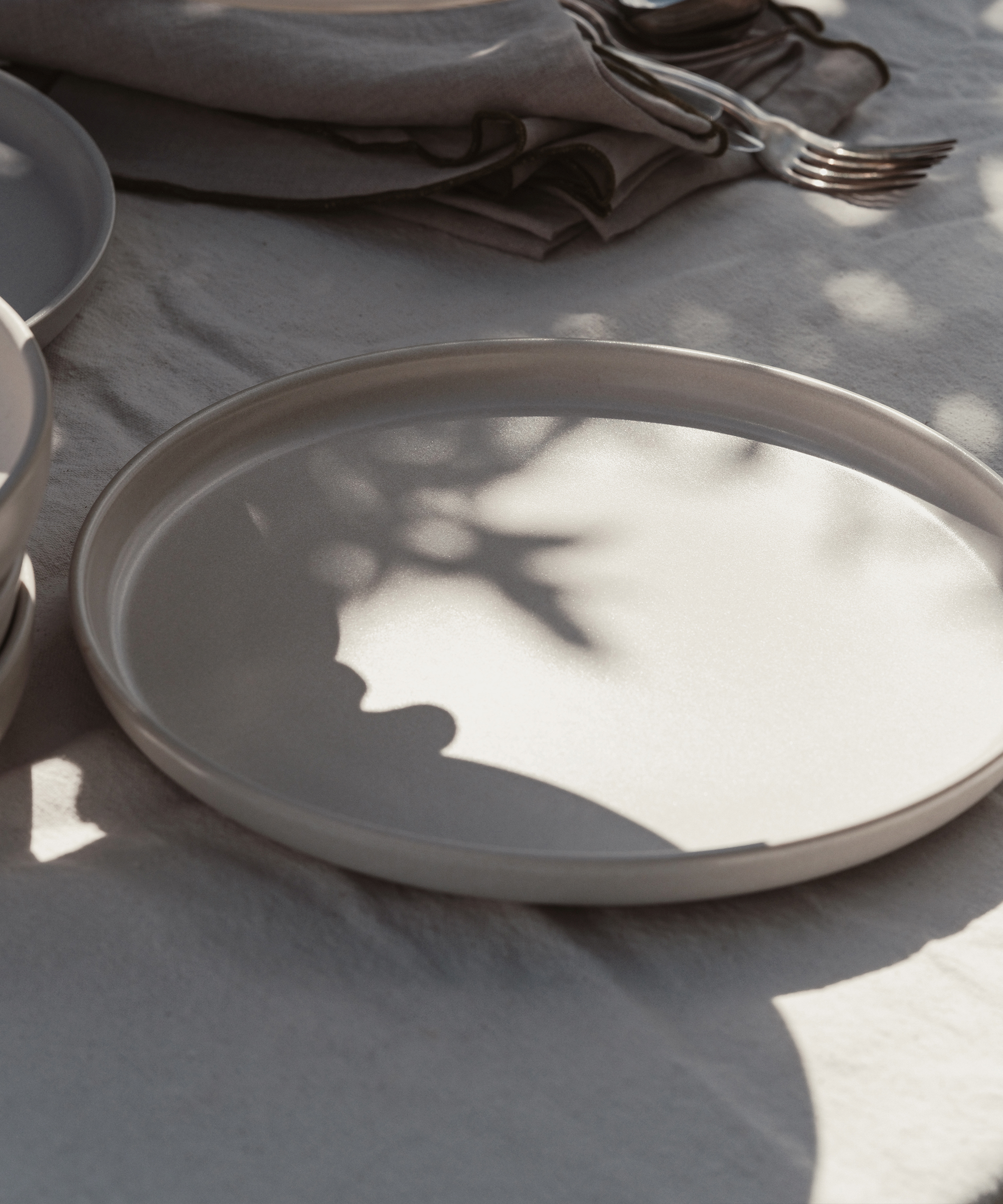 A plain, round, beige plate sits on a tablecloth with soft sunlight and leafy shadows. Folded napkins and forks in the background evoke a calm, minimalist table setting inspired by Michèle Ouellet Benson’s understated elegance.