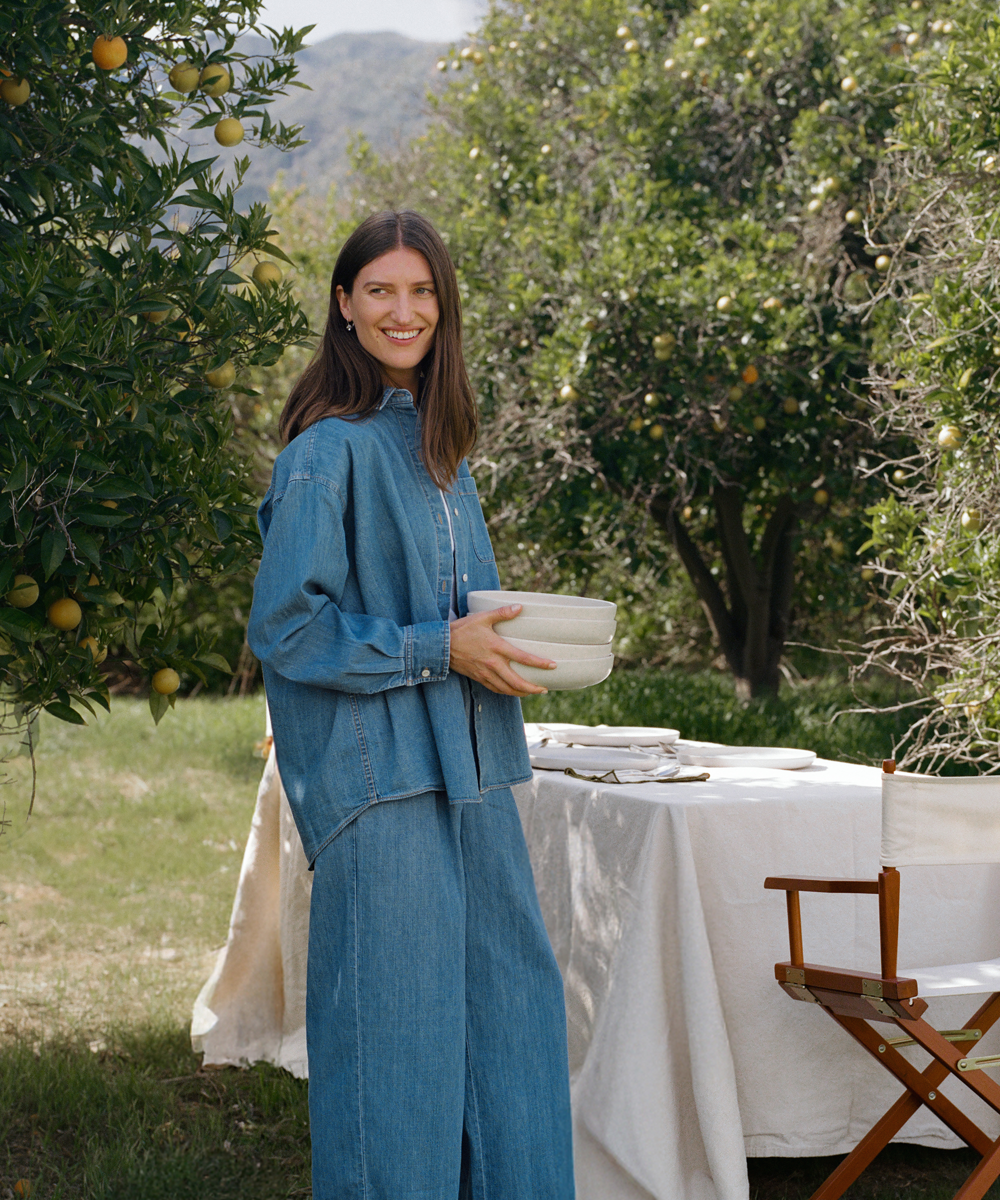 Michèle Ouellet Benson stands outdoors in a denim shirt and jeans, smiling as she holds stacked bowls near a table set for a meal in a lush garden with citrus trees and mountains in the background.