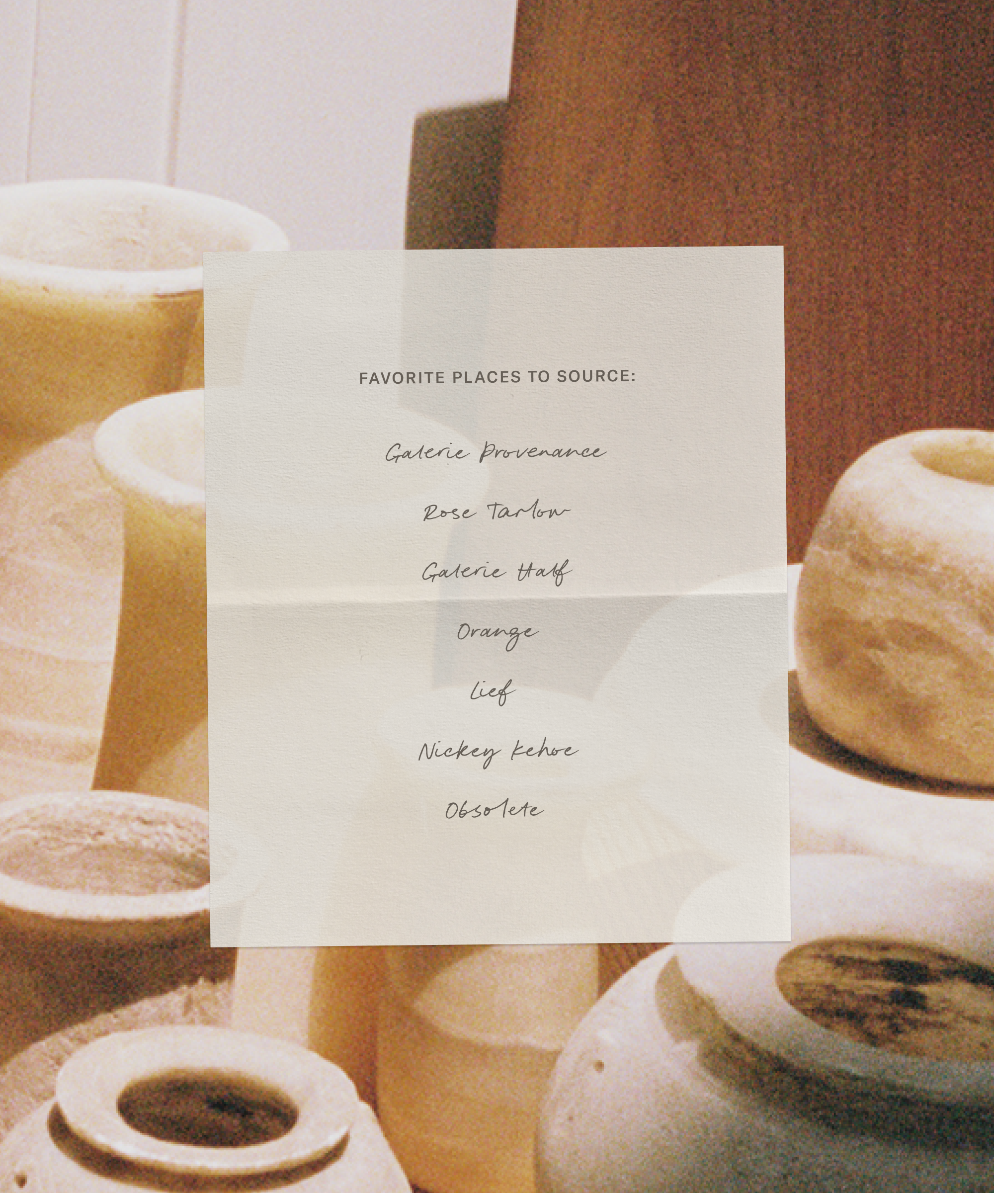 A paper with a handwritten list of favorite places to source is taped to a surface. In the background are several cream-colored ceramic vases and vessels, softly lit.