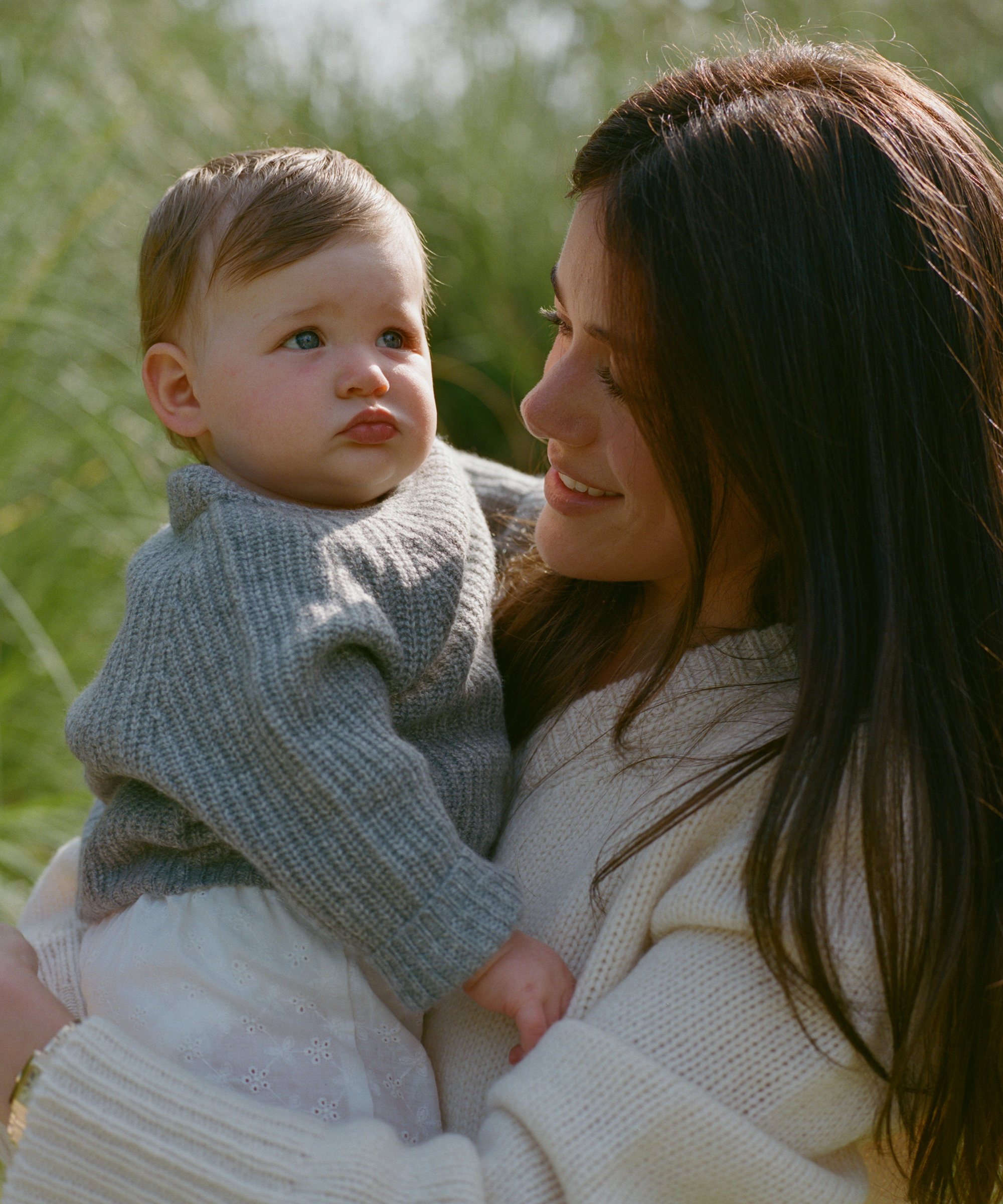 a mom in an ivory cashmere sweater holding her daughter up in a grey cashmere sweater outside in a park
