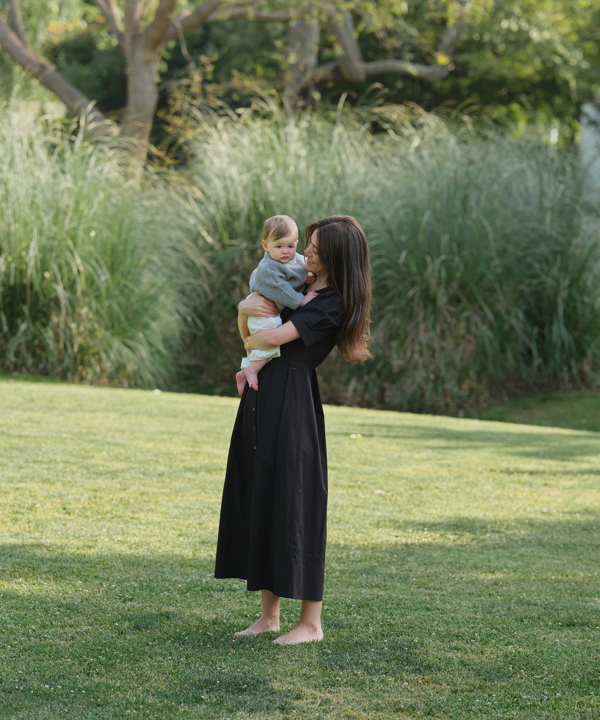 Ilana Torbiner captures a woman in a long black dress standing barefoot on grass, holding a small child. They are outdoors in a park, surrounded by tall green grass and trees in the background.