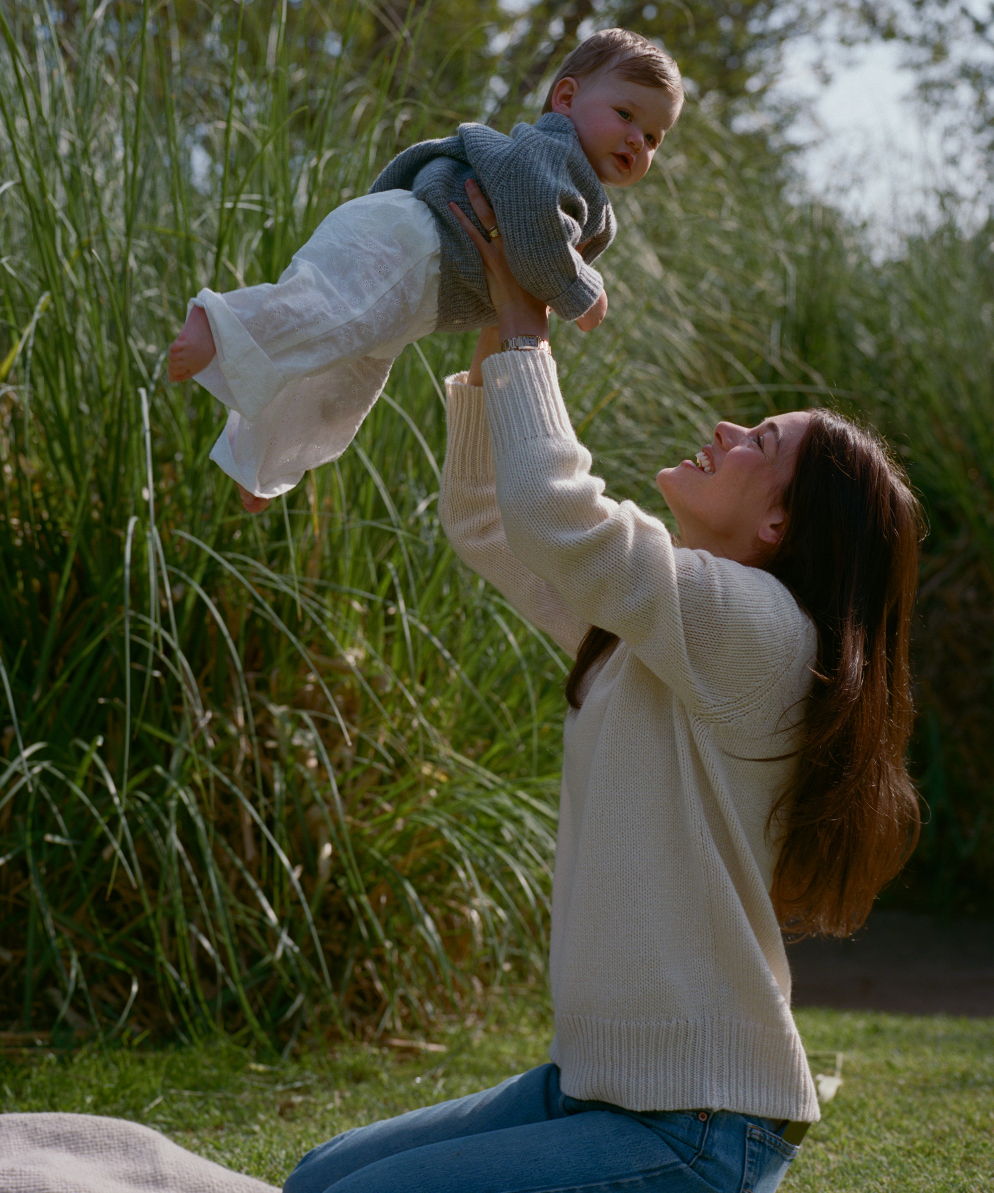 a mom in an ivory cashmere sweater holding her daughter up in a grey cashmere sweater outside in a park