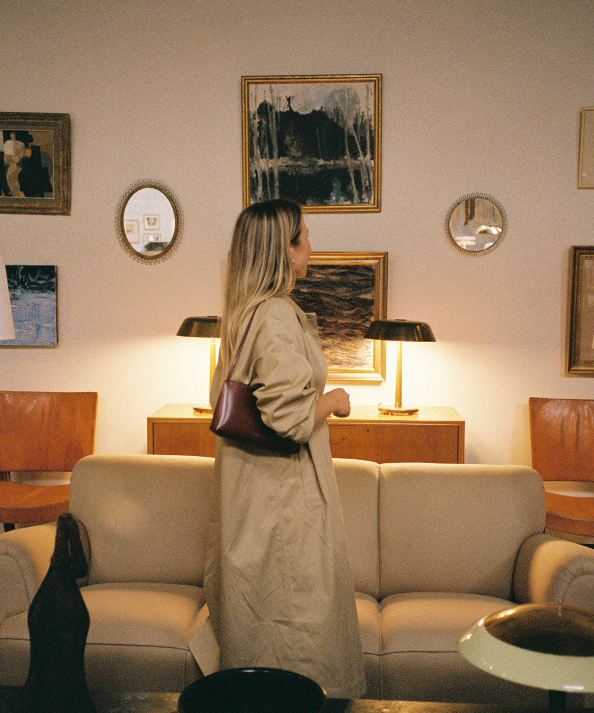 A woman in a beige trench coat stands in a stylish living room, facing a wall decorated with framed artwork and mirrors above a beige sofa and wooden sideboard with two lamps.