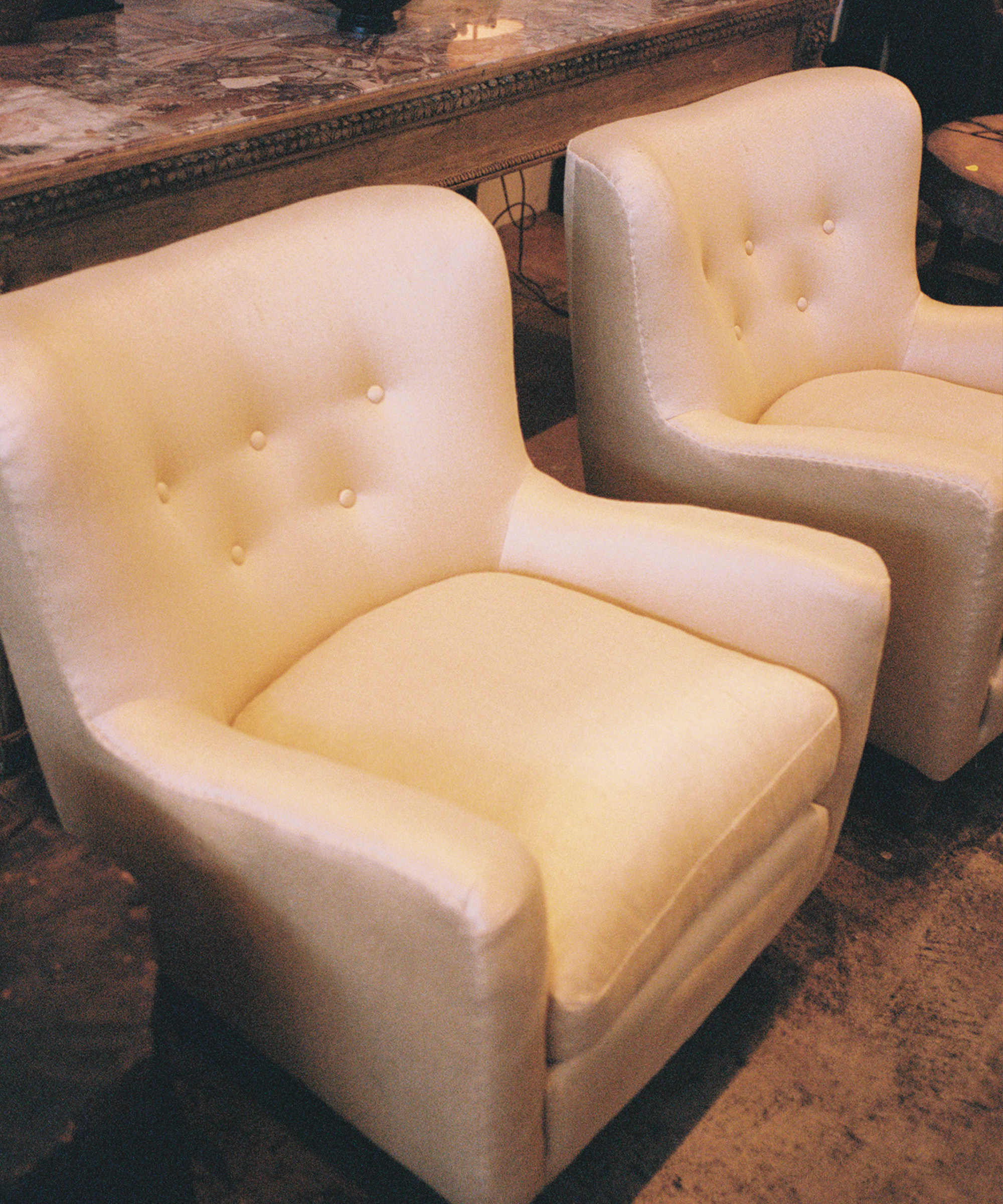 Two cream-colored, upholstered armchairs with button tufting on the backrests are placed side by side on a rustic floor, in front of a marble-top wooden table.