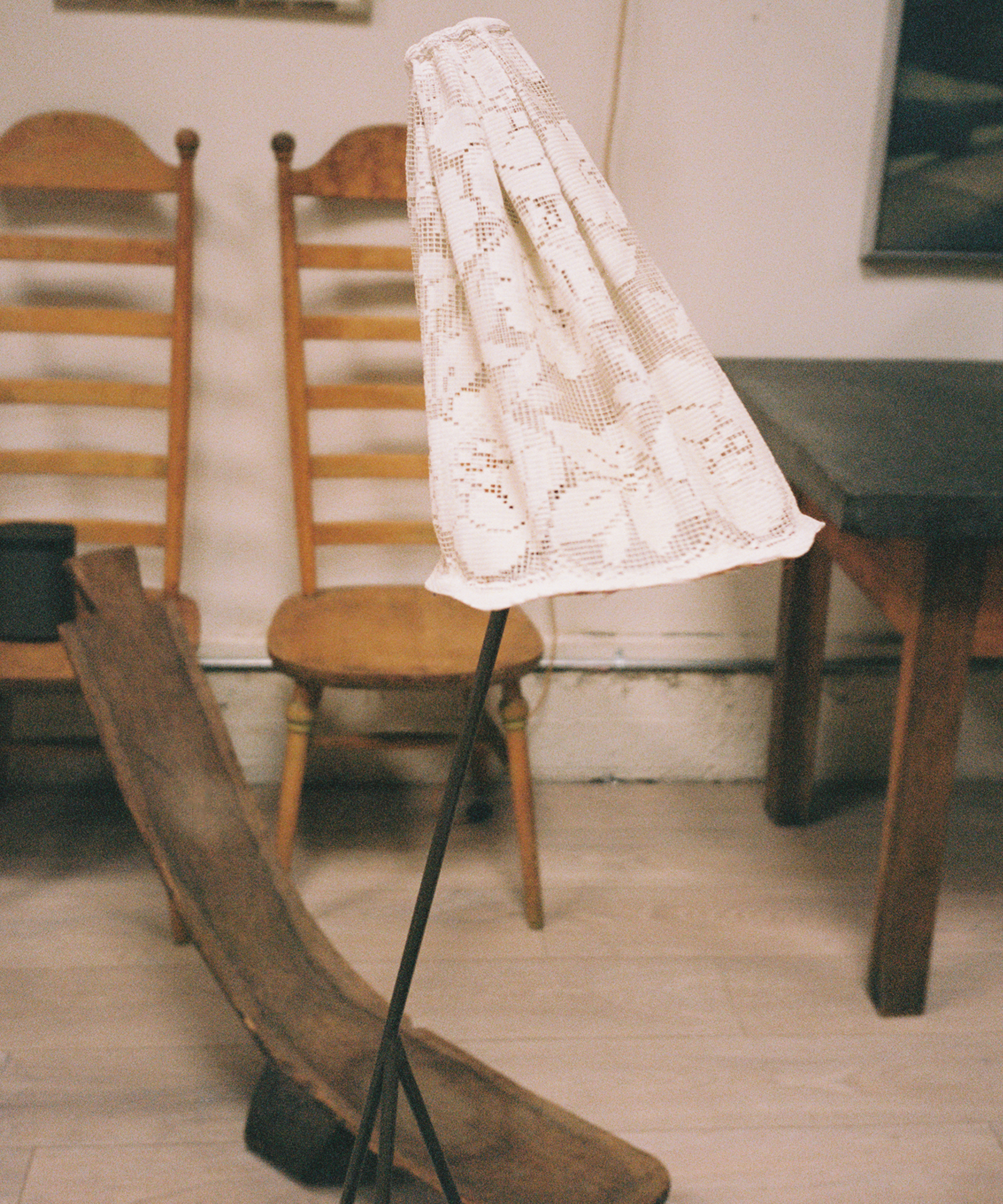 A standing lamp with a lace-covered shade is in front of two wooden chairs and a rustic wooden trough, with a dark table to the right, all set in a room with light wood flooring.