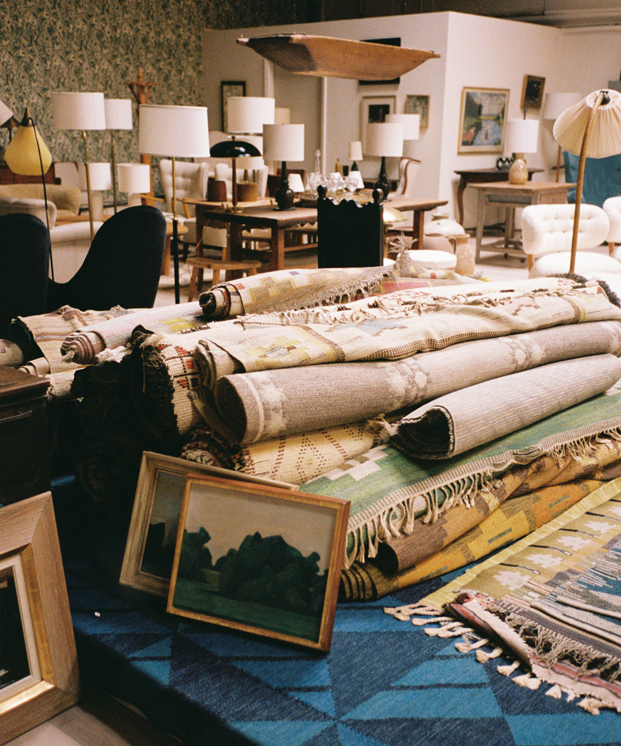 A stack of rolled rugs sits on a blue patterned carpet, surrounded by framed artwork and various lamps, tables, and chairs in a vintage shop setting.