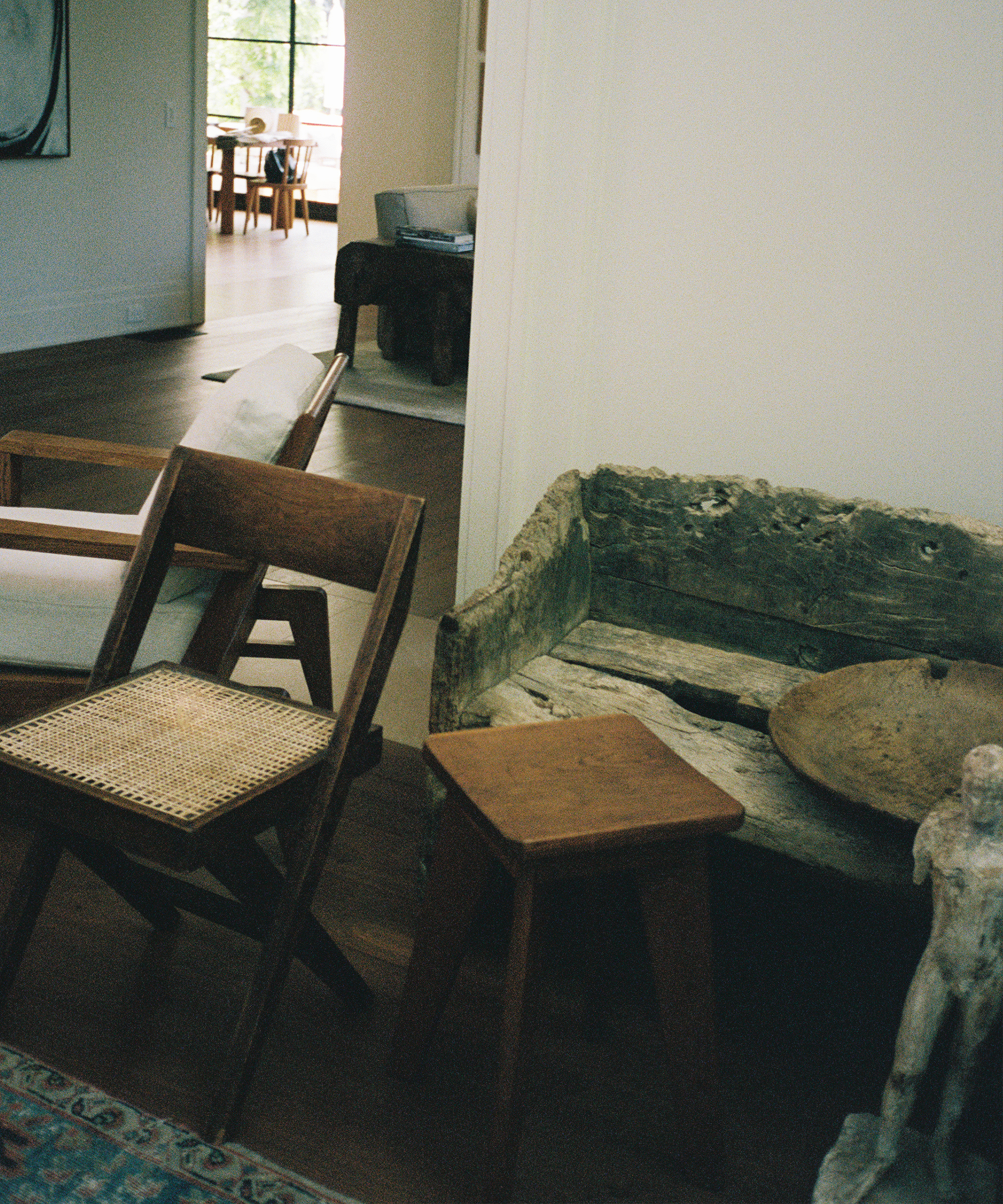 A cozy interior with wooden furniture, including a rustic bench, two wooden chairs, a small stool, and a sculpted figure, set on a wooden floor near a sunlit room with large windows in the background.
