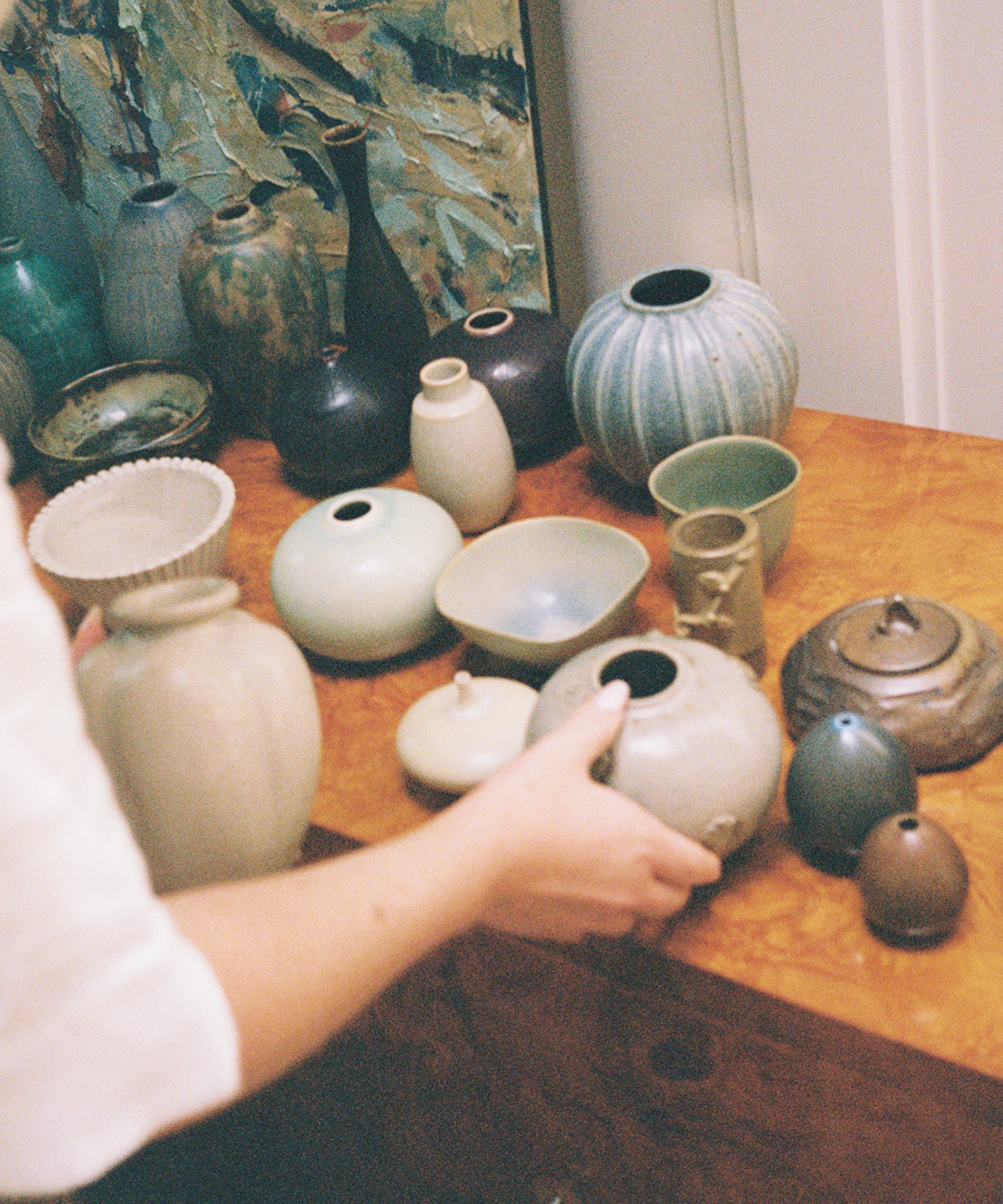 A person arranges various ceramic vases, bowls, and pots in muted earth tones on a wooden surface, with abstract artwork and more pottery in the background.