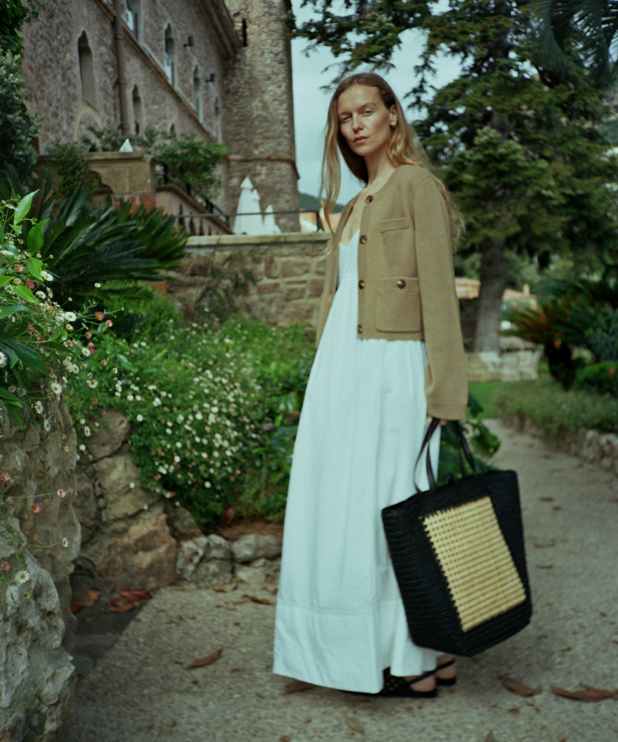A woman in a long white dress and tan jacket stands outdoors on a pathway, holding a black woven tote bag. Channeling the effortless style of Marie von Behrens-Felipe, she is surrounded by greenery, stone walls, and a large tree.