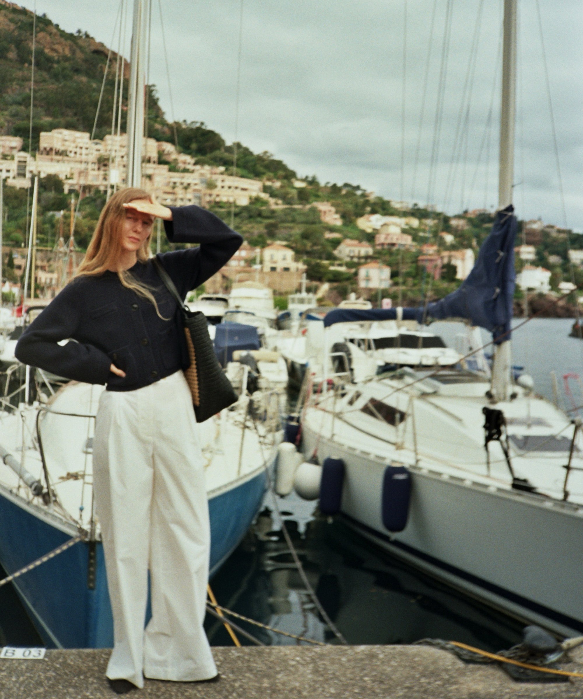 A woman, reminiscent of Marie von Behrens-Felipe, stands by a marina in white pants and a dark top, shielding her eyes from the sun. Behind her, sailboats line the docks with hills and houses under a cloudy sky.
