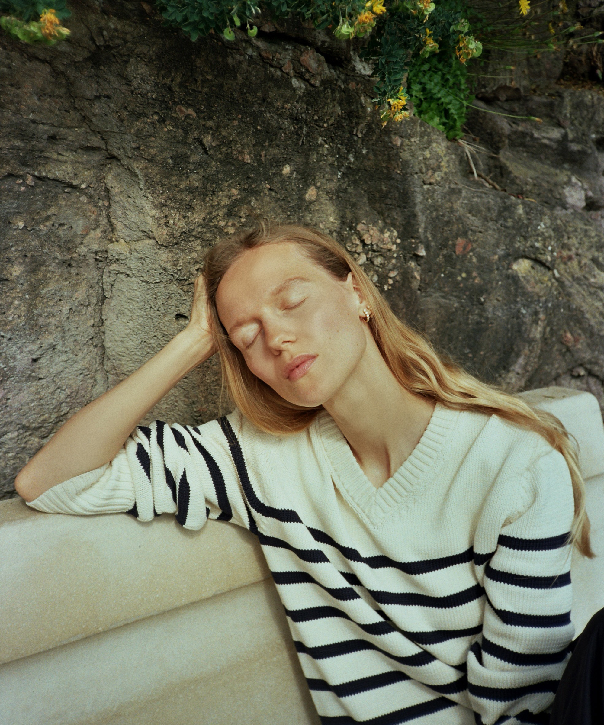Marie von Behrens-Felipe, with her signature long blonde hair, sits on a bench in a white and navy striped sweater, eyes closed and head resting on her hand, against a stone wall framed by lush greenery.