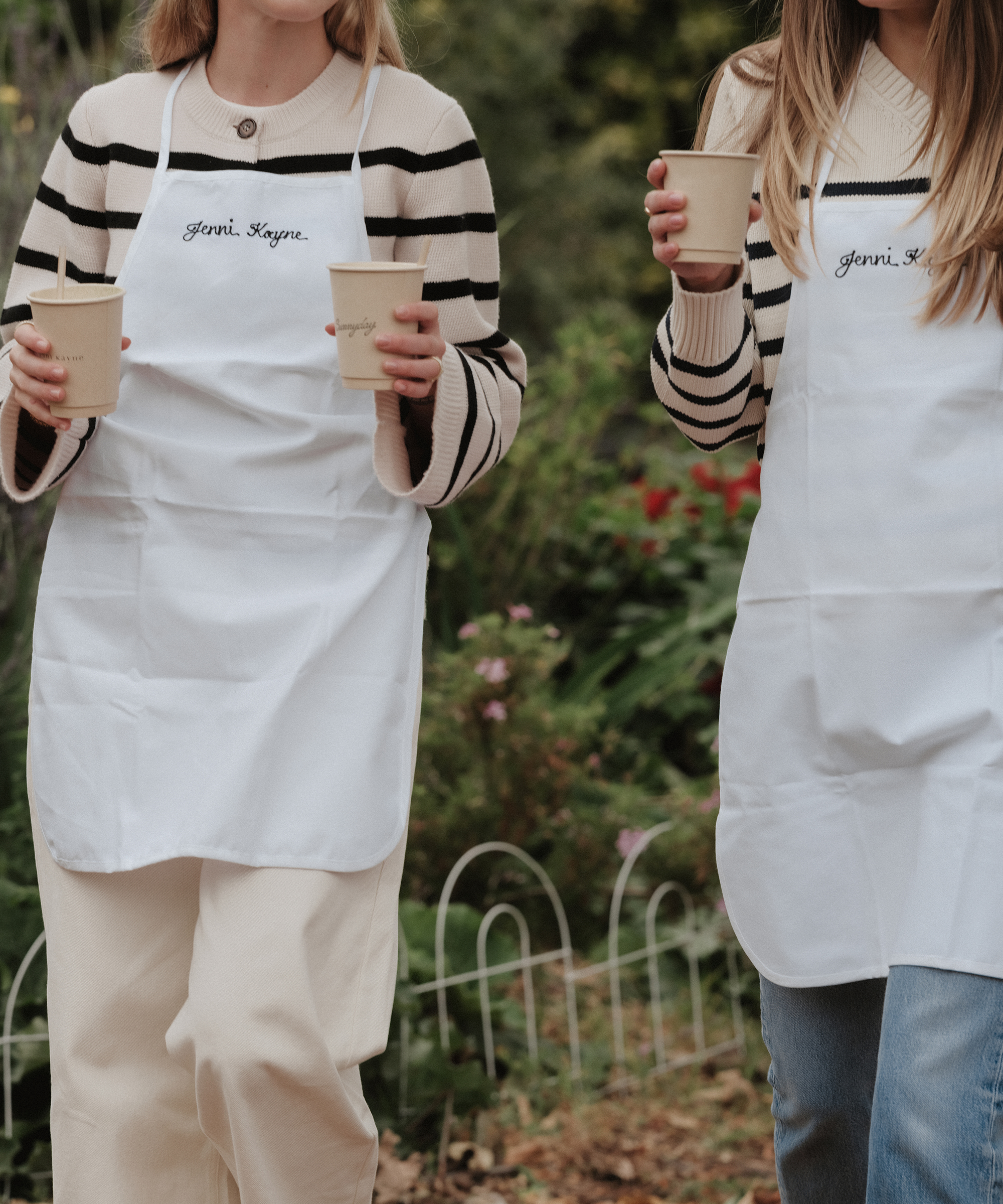 Two women wearing white aprons and striped sweaters hold paper cups while standing outdoors in a garden at Thorne Family Farms. Their faces are not visible in the image.
