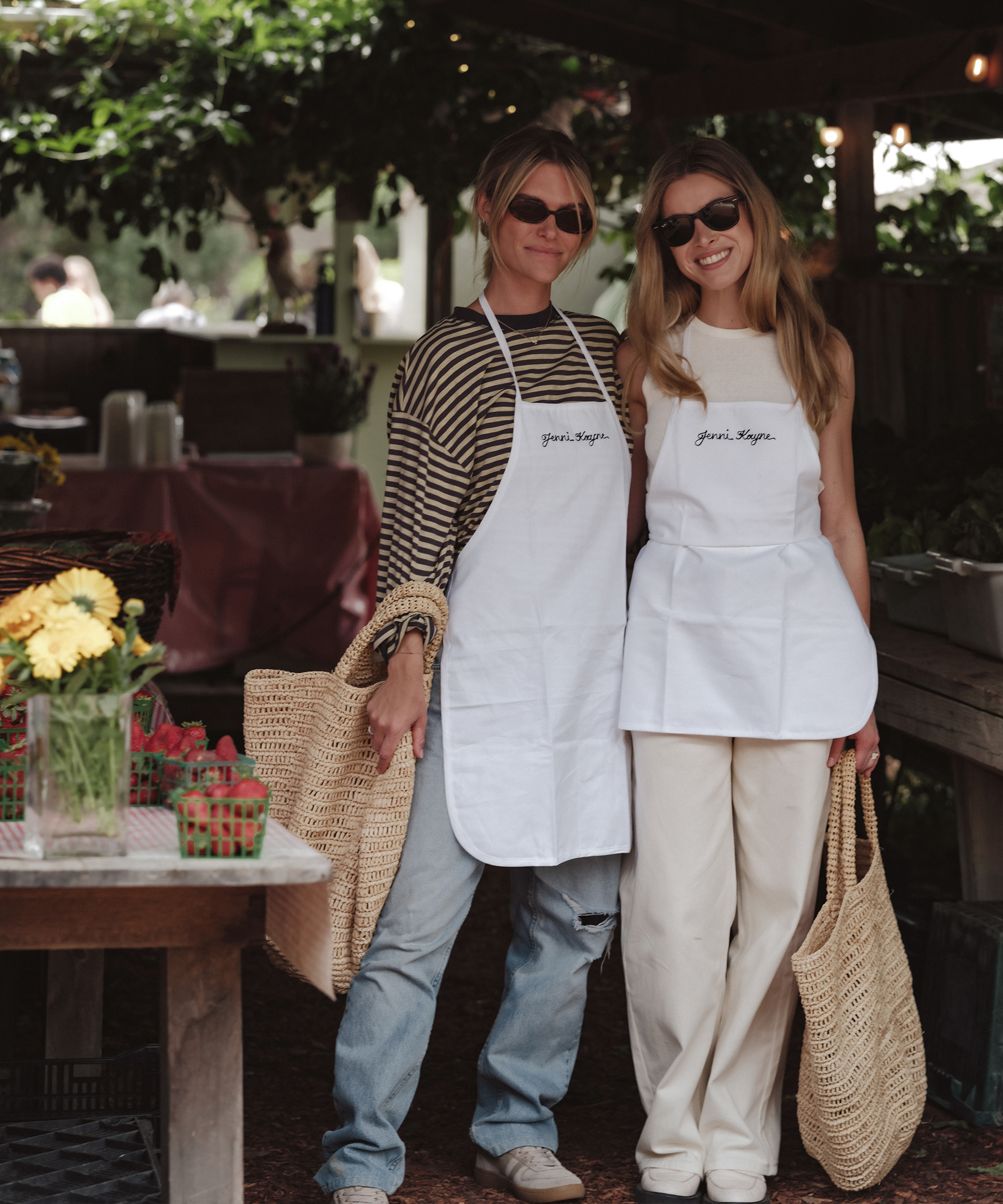 two girls wearing white linen aprons with jenni kayne embroidered and jeans inside a market holding crochet raffia totes