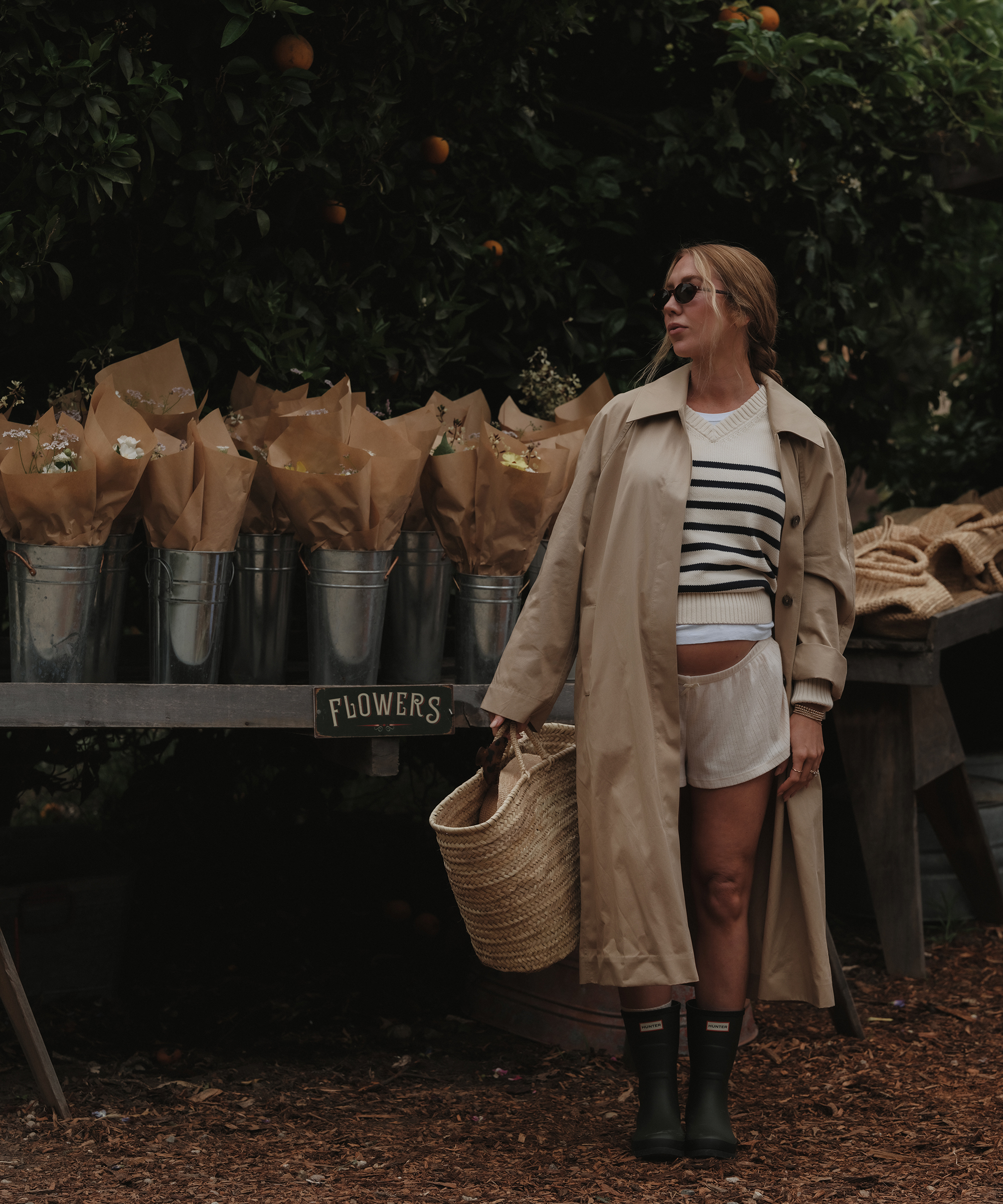 A woman in a trench coat, striped shirt, and rain boots stands by a table of flower bouquets from Thorne Family Farms, wrapped in brown paper, holding a straw basket in an outdoor market setting with greenery in the background.