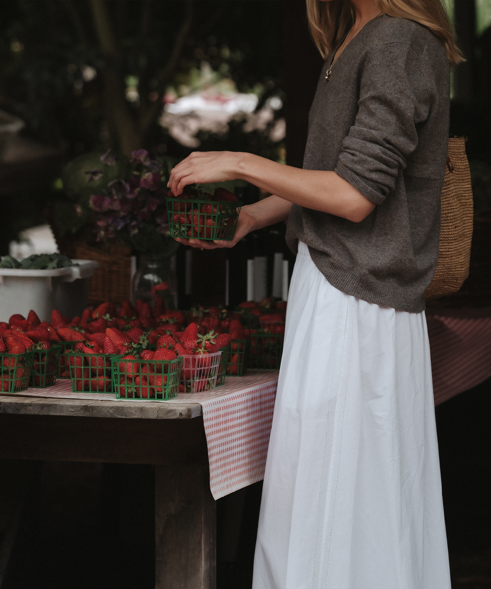 A woman in a gray sweater and white skirt picks up a basket of strawberries at the Thorne Family Farms outdoor market stand, where several baskets of fresh strawberries are displayed on a table.