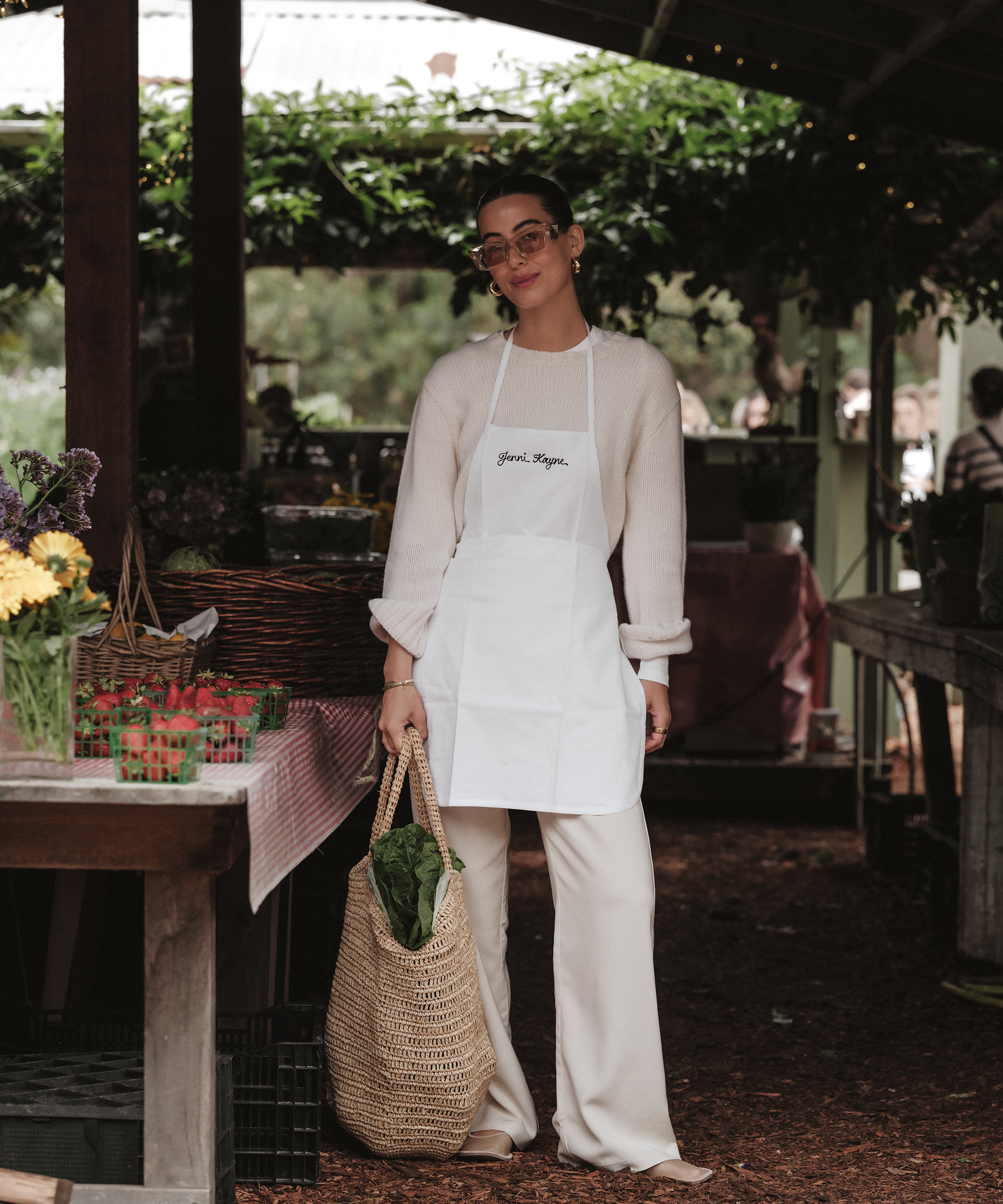 A woman wearing glasses, a white apron, cream sweater, and pants stands at a Thorne Family Farms outdoor market stall holding a woven bag with greens. Flowers, strawberries, and baskets are visible on the tables around her.