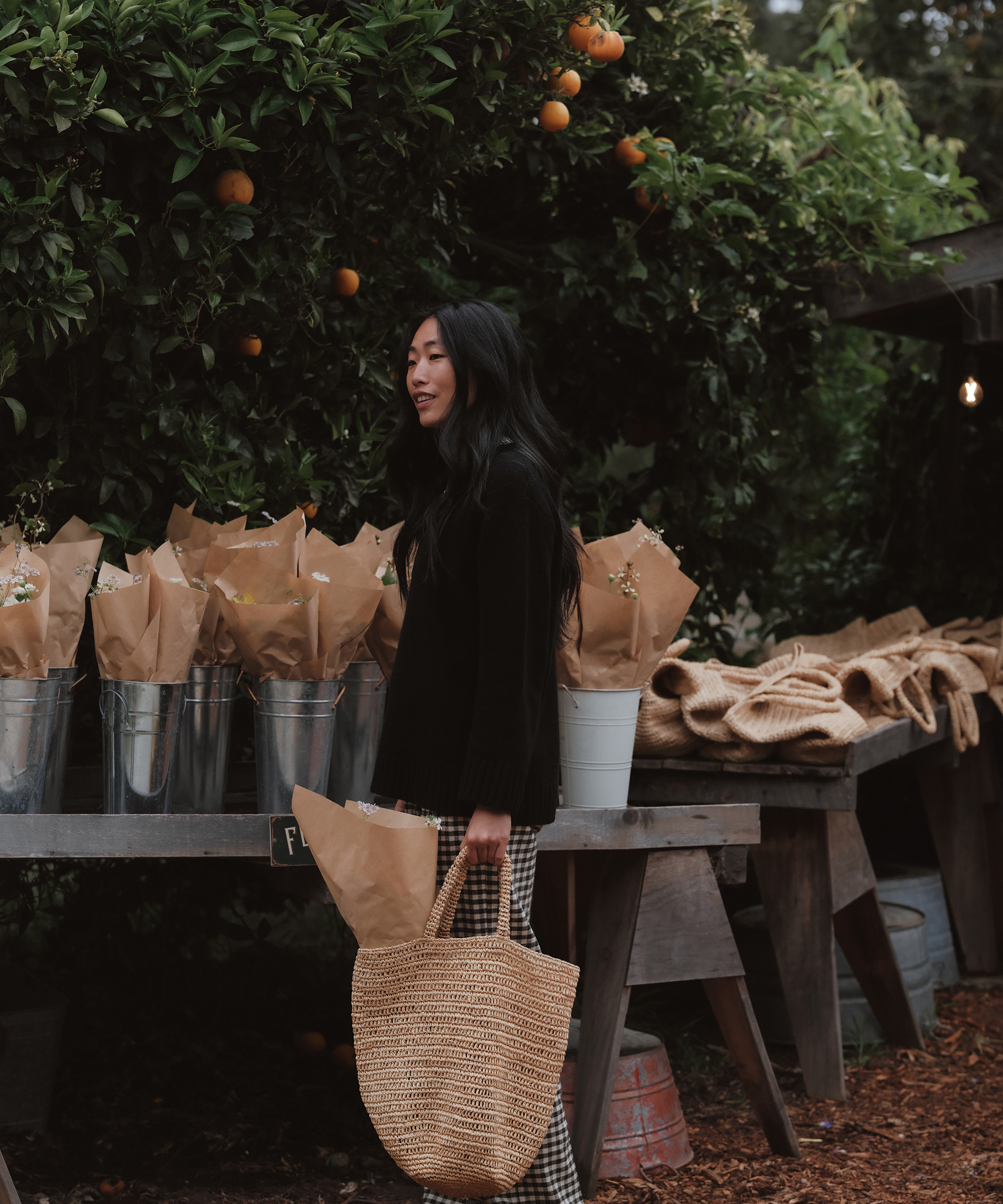 a woman in a black cashmere sweater and plain pants holding a raffia tote selecting florals from buckets outside a farmers market