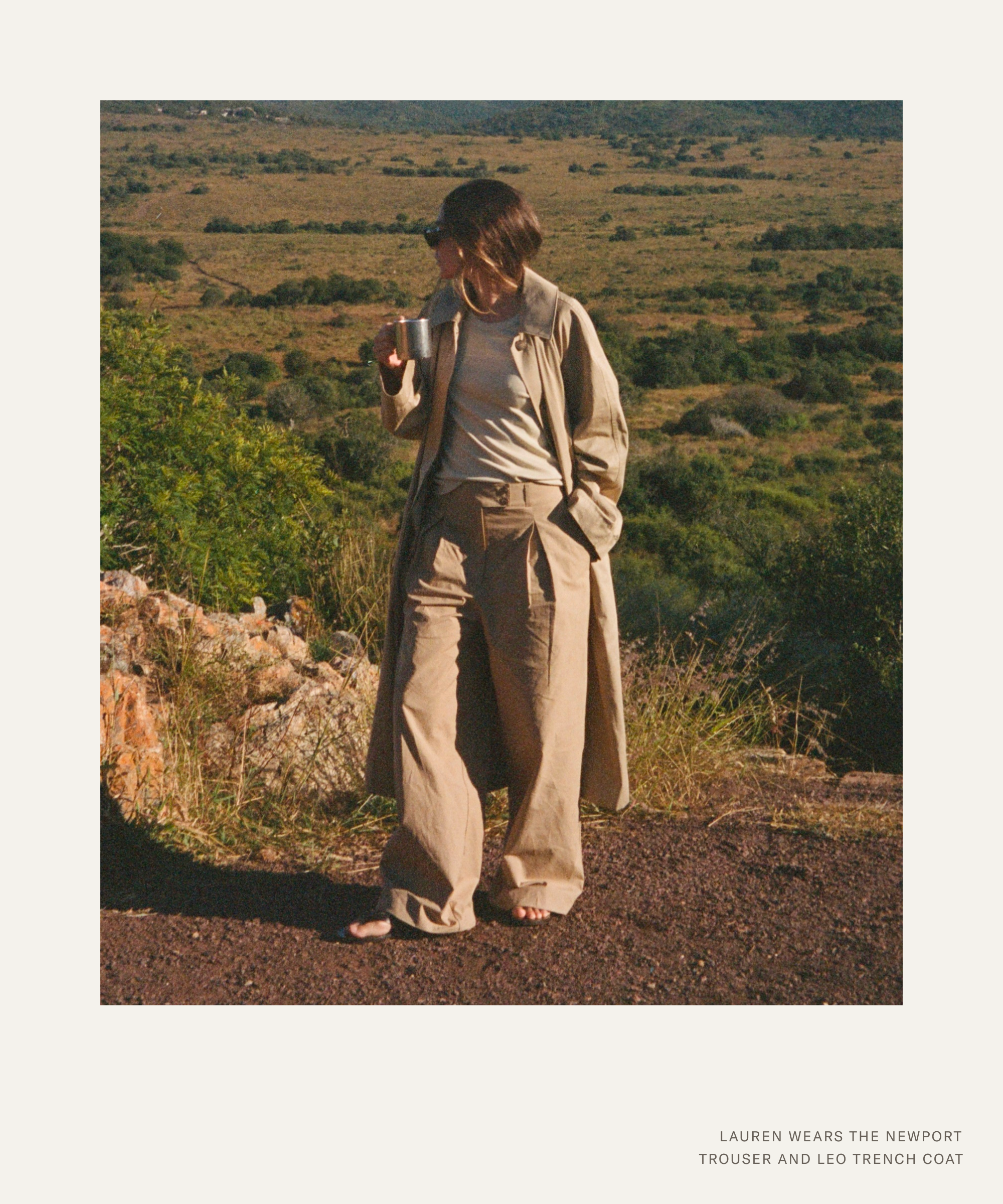 A person stands outdoors on a dirt path, wearing a beige trench coat and matching trousers, looking to the side while holding a cup, with a green landscape stretching out behind them—a scene evoking the effortless style of Lauren Burke.