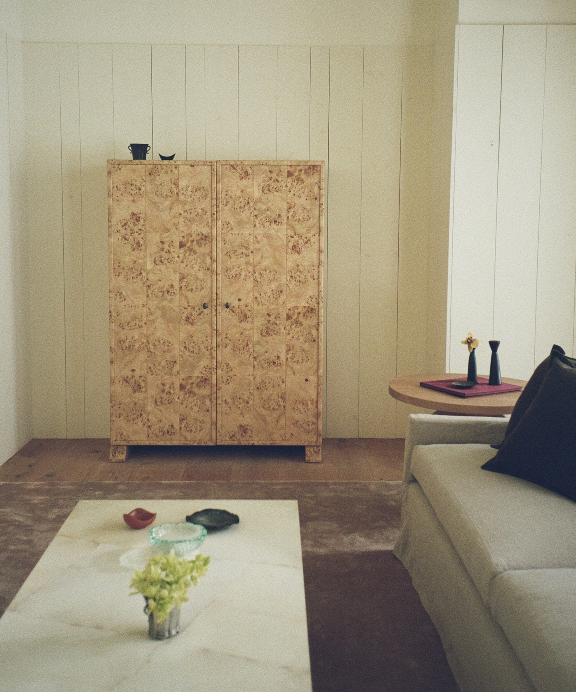 A minimalist living room with a beige sofa, a white marble coffee table with a small floral arrangement, a light wood cabinet, and a round side table holding two vases. The room features light-colored paneled walls.