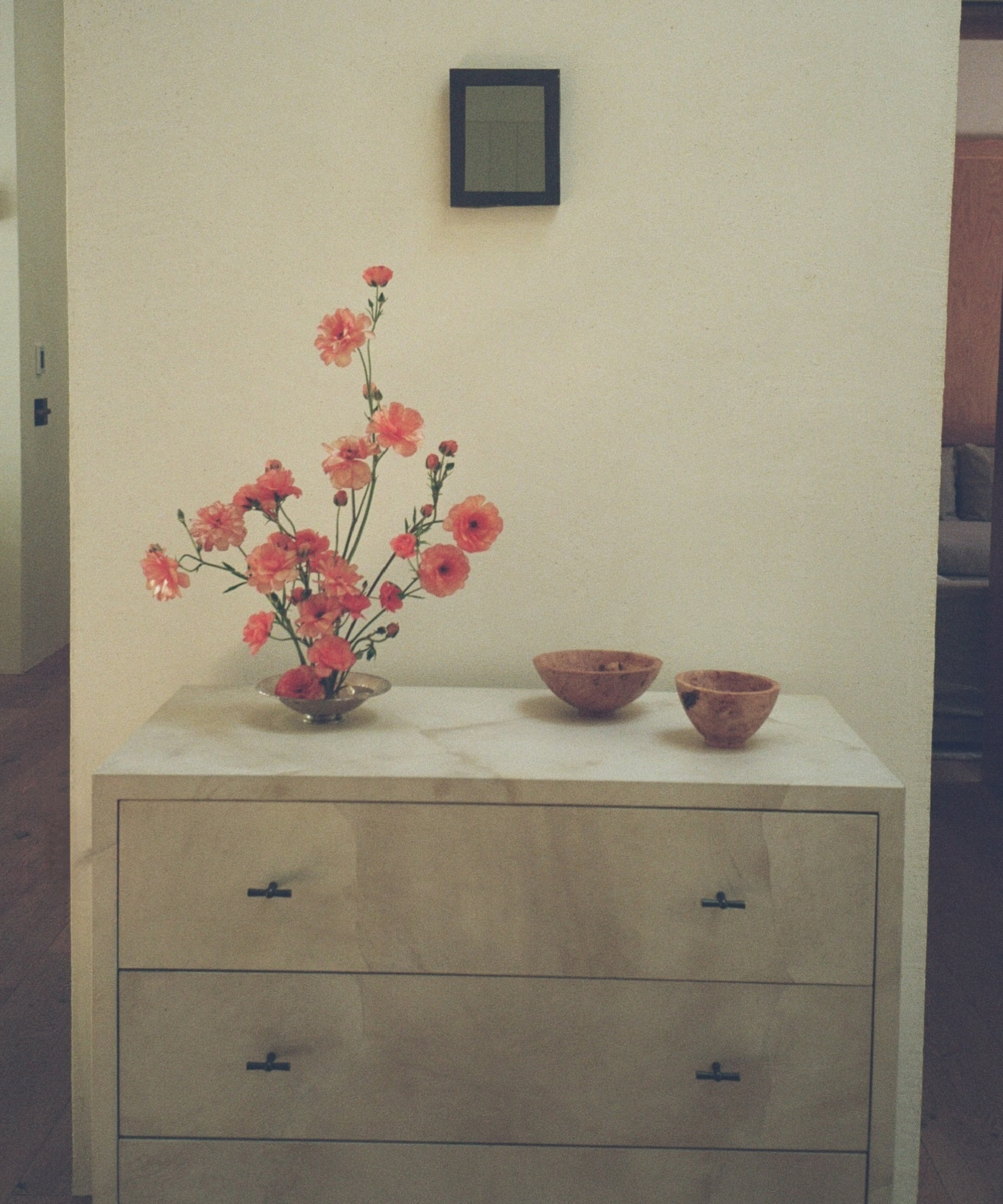 A beige dresser with three drawers holds a glass vase of pink flowers and two wooden bowls. A small, black-framed square picture hangs on the cream-colored wall above the dresser.