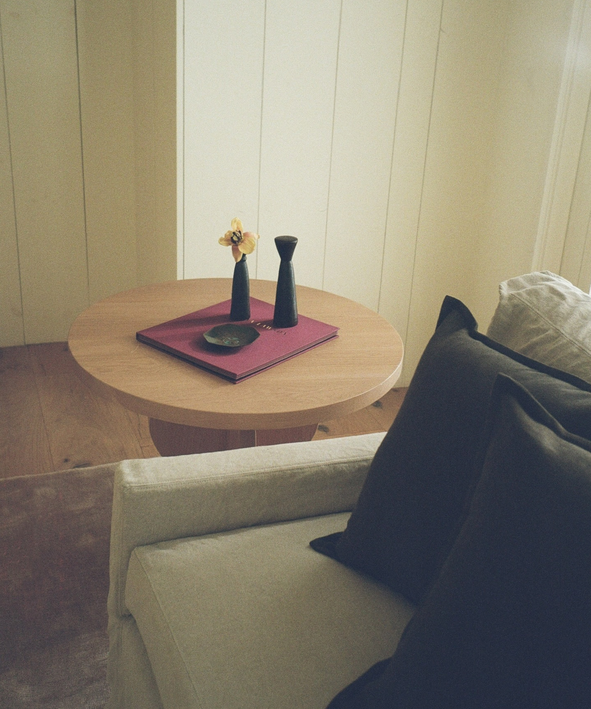 A round wooden side table holds a purple tray with two black vases, one containing a yellow flower, beside a light-colored sofa with dark cushions in a softly lit room.