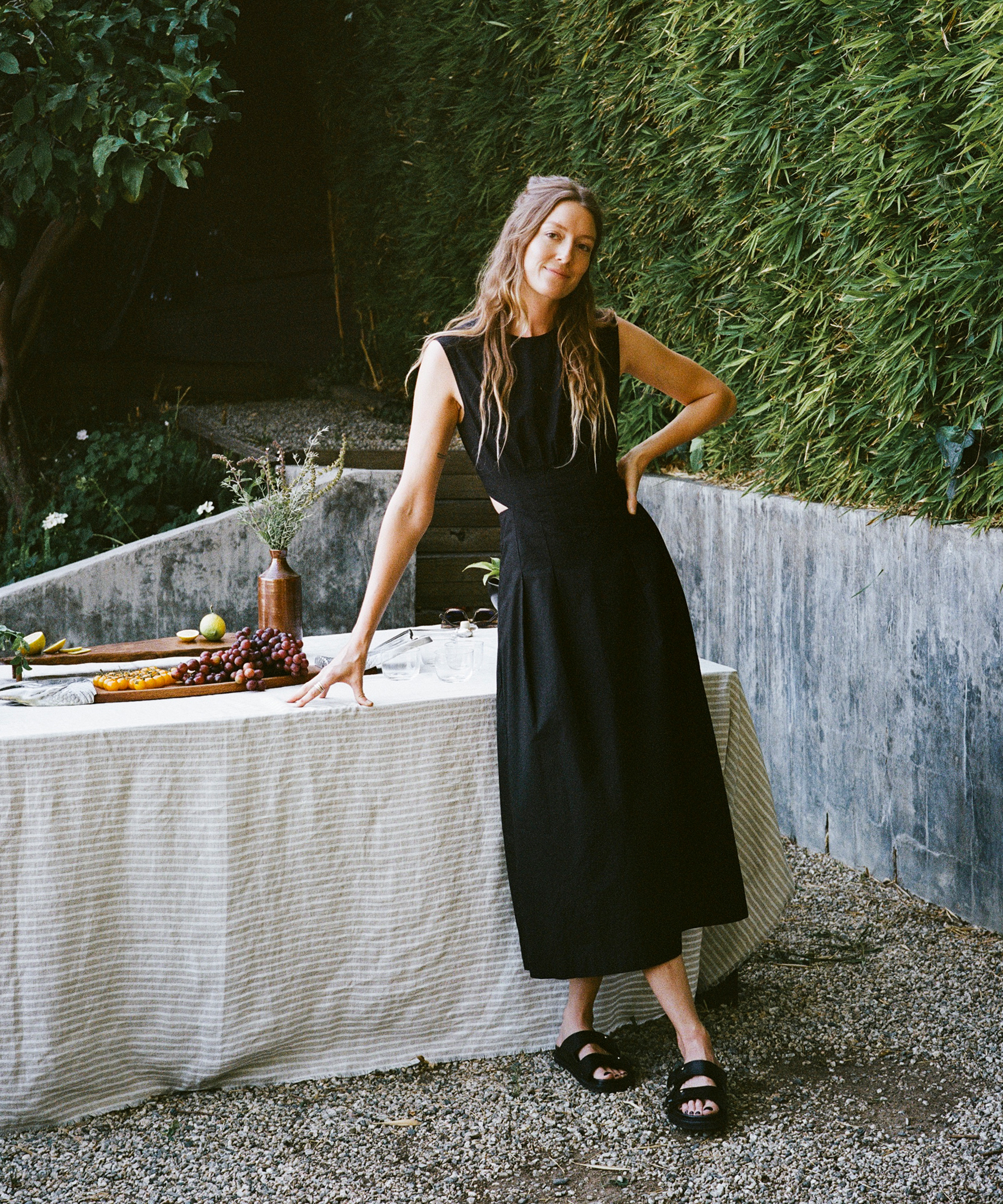 a woman wearing a black maxi dress and black sandals standing next to a striped linen tablecloth