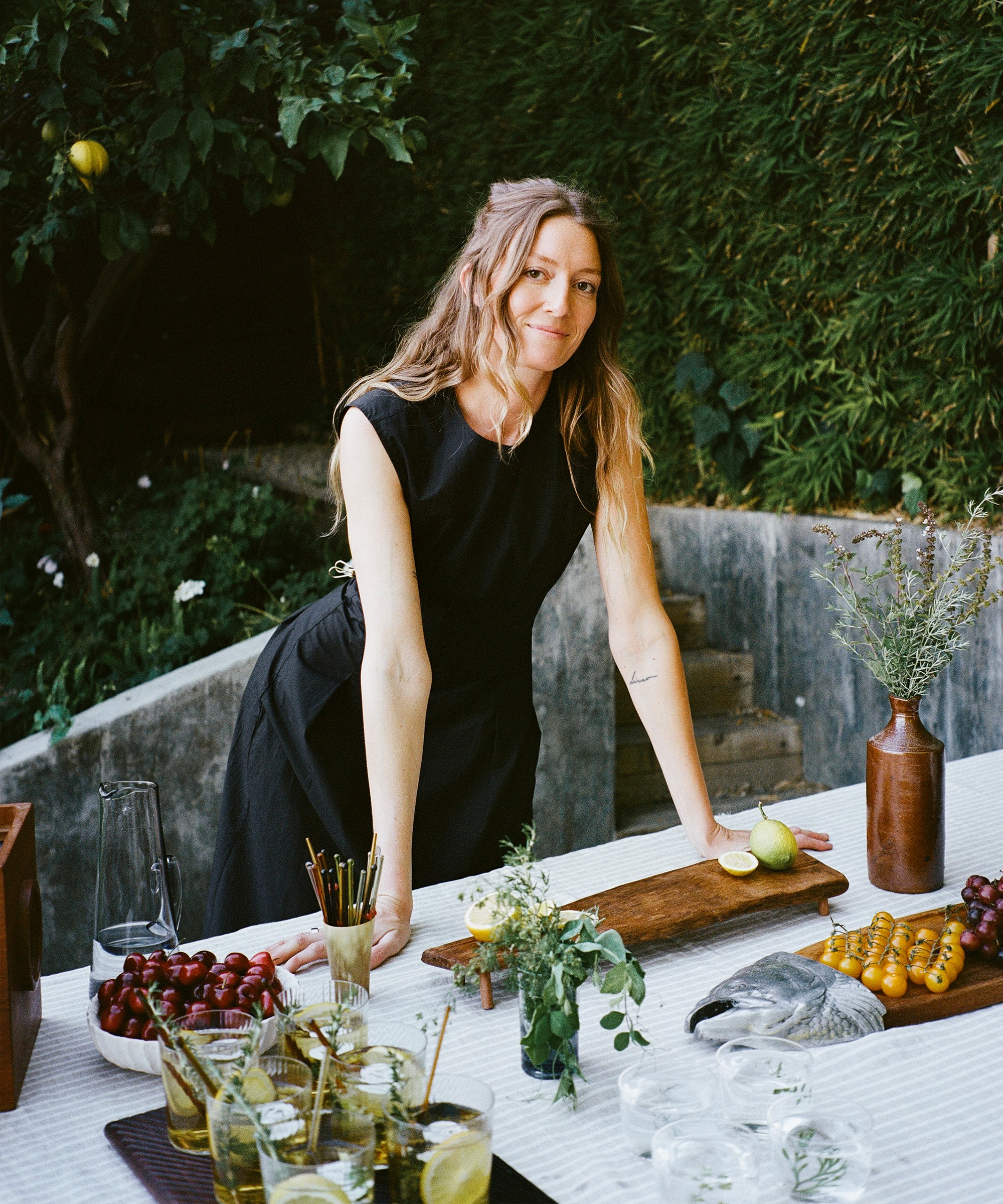 a woman wearing a black dress leaning against an outdoor dining table filled with foods and drinks