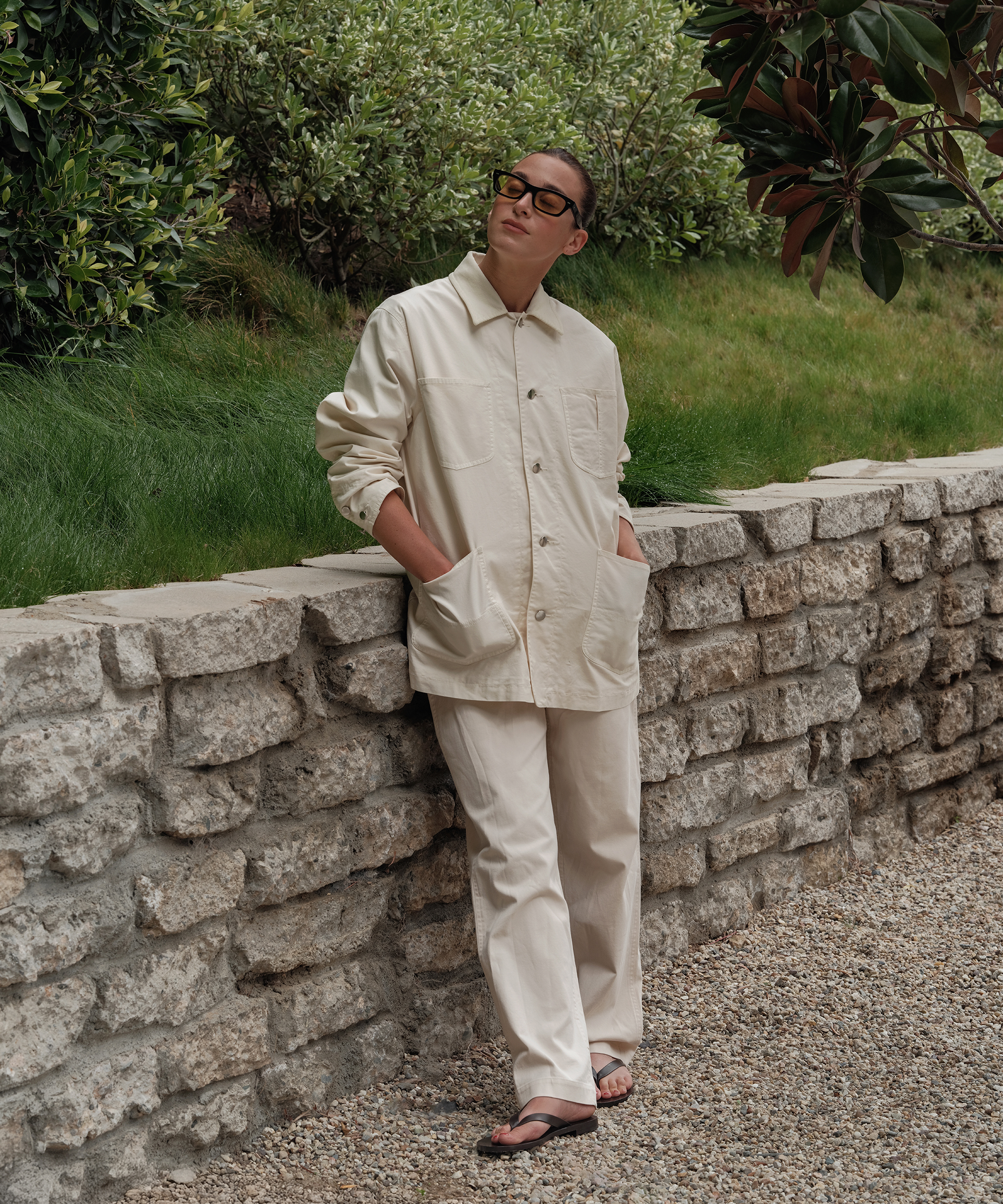 A person wearing cream-colored pants and a matching button-up jacket stands against a stone wall, hands in pockets, wearing sunglasses and sandals, with greenery in the background.