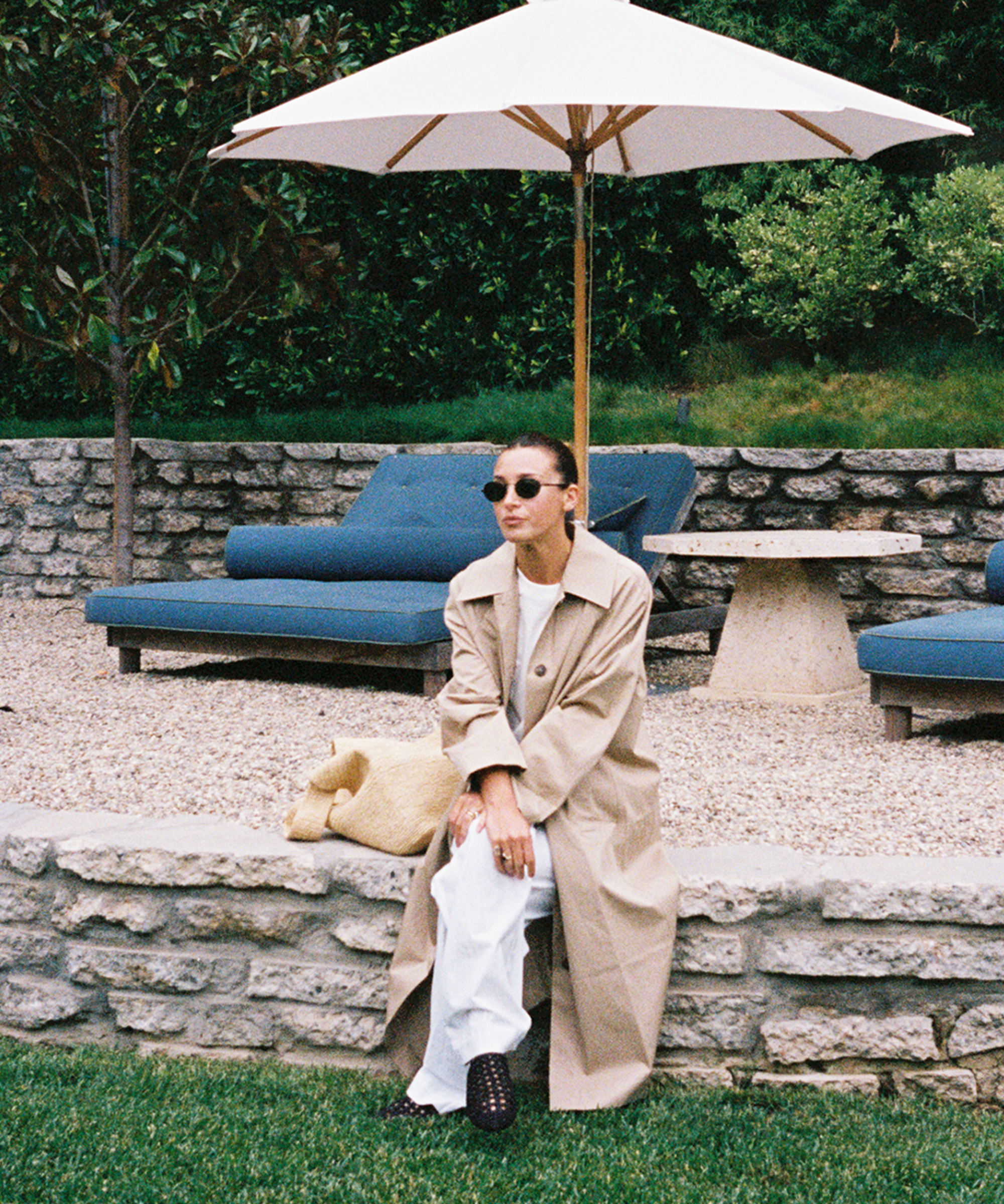 A woman in sunglasses and a long beige trench coat sits on a stone ledge under a white patio umbrella, with blue outdoor lounge chairs and greenery in the background.