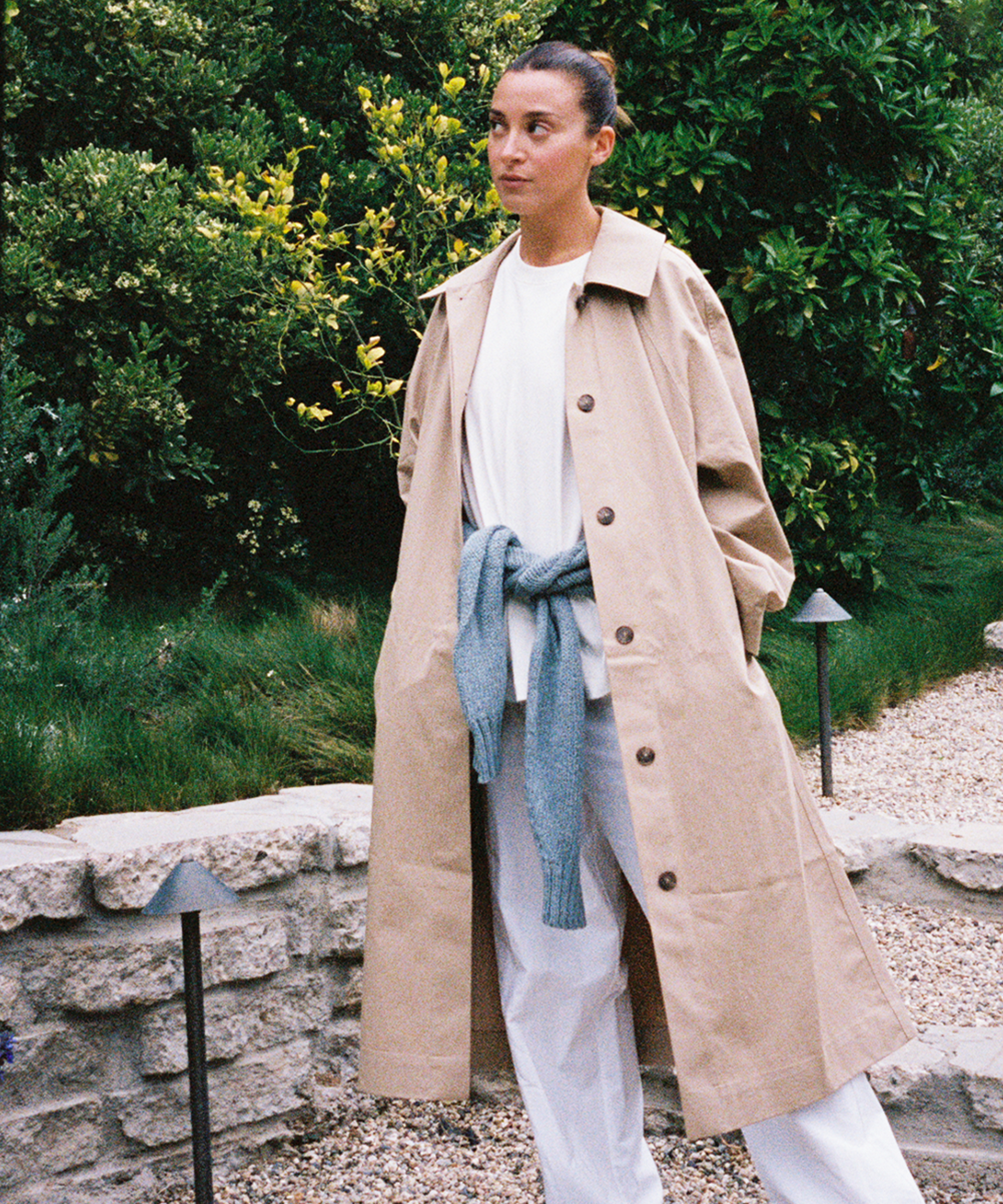 A woman stands outdoors in front of greenery, wearing a long beige trench coat over a white outfit. A light blue sweater is tied around her waist, and she looks off to the side with her hands in her coat pockets.