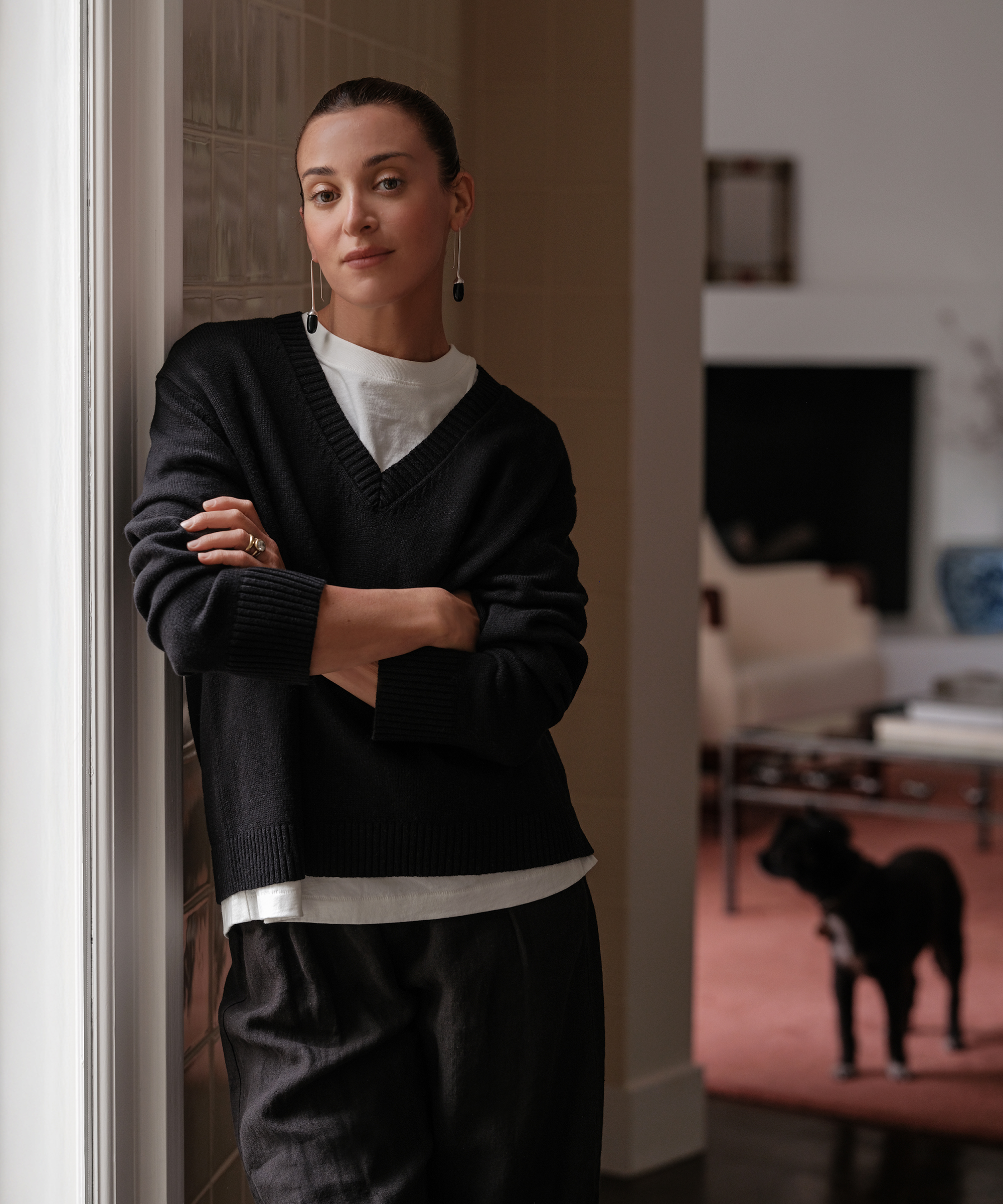 A woman with her arms crossed leans against a wall in a softly lit room, wearing a black sweater over a white shirt. A black dog stands in the background near a pink rug and modern furniture.