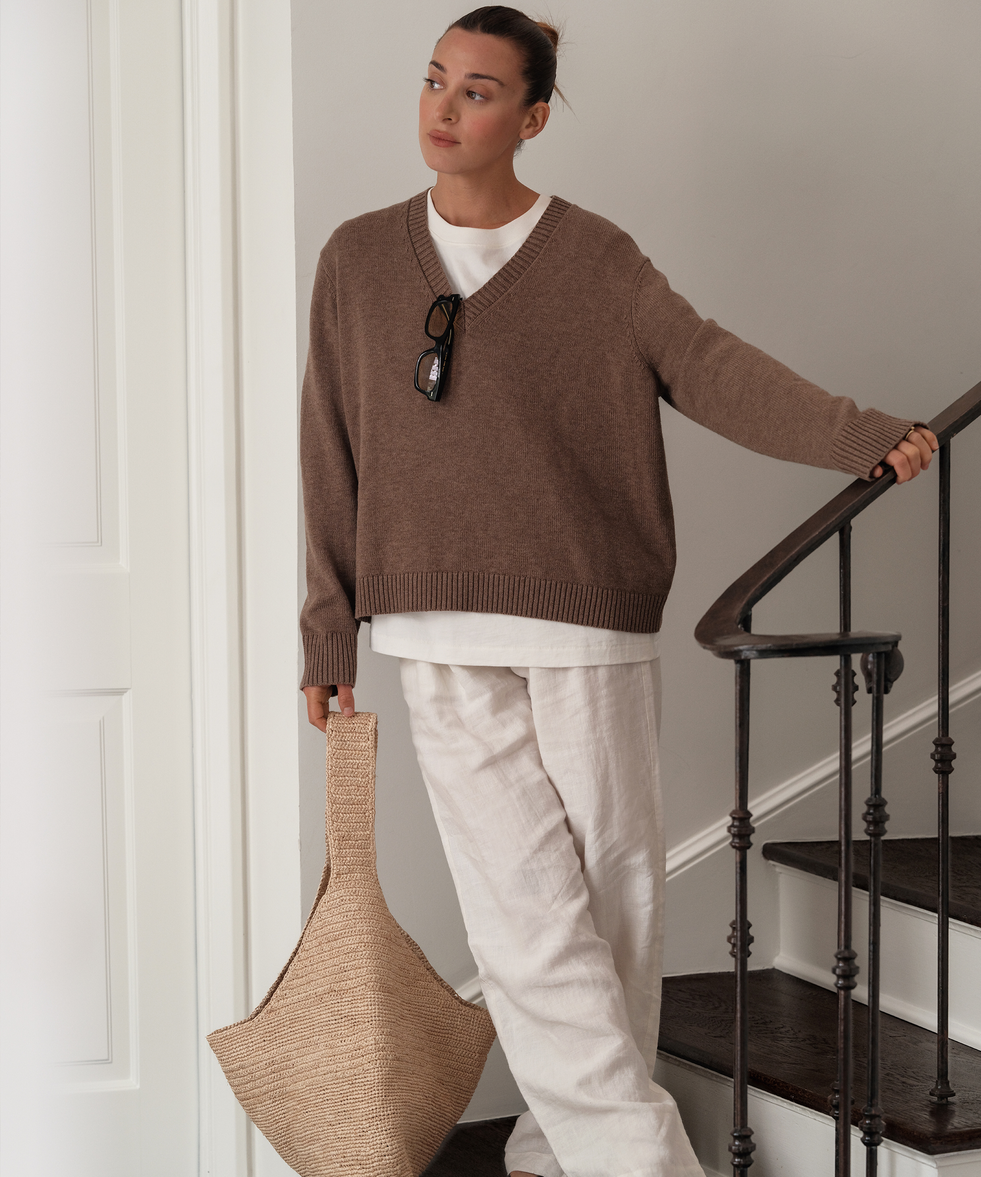 A woman stands on a staircase holding the railing and a woven straw bag. She wears a brown oversized sweater over a white shirt, loose beige linen pants, and has sunglasses hanging from her sweater’s neckline.