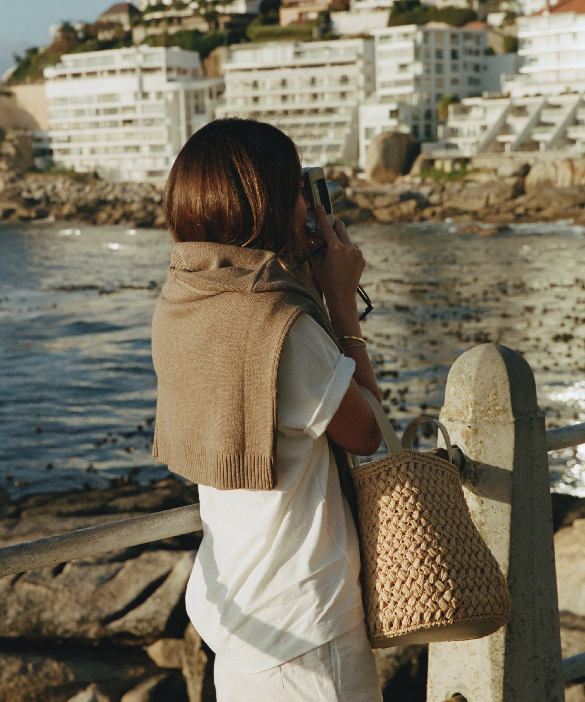 a woman wearing a white tee with a doe colored sweater over her shoulders taking a photo by the beach