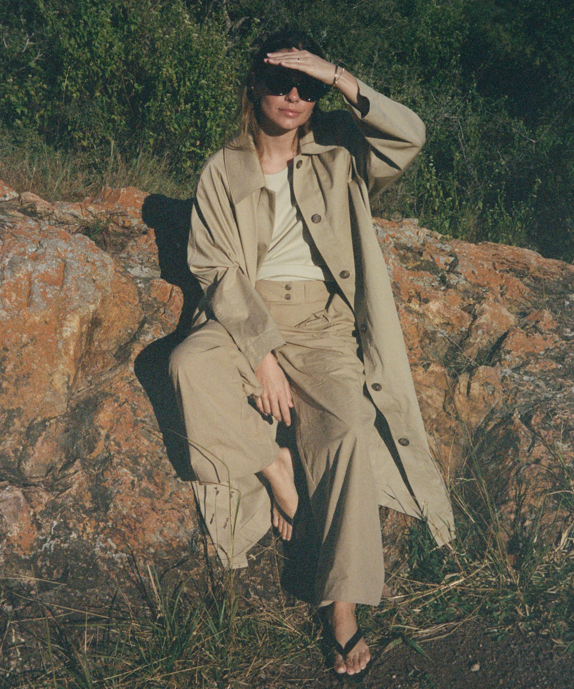 a woman sitting on a rock wearing a khaki trench and khaki trousers