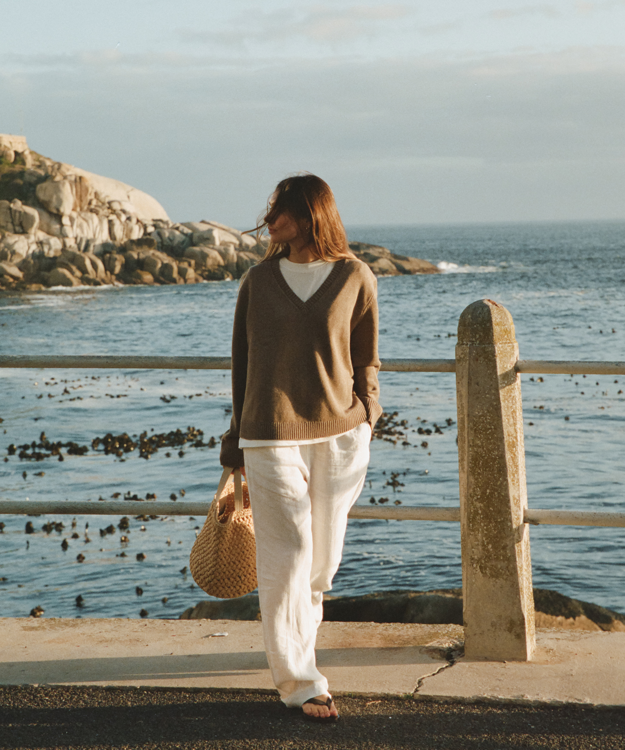 lauren wearing a doe colored light sweater ivory linen pants and a raffia tote standing in front of the ocean
