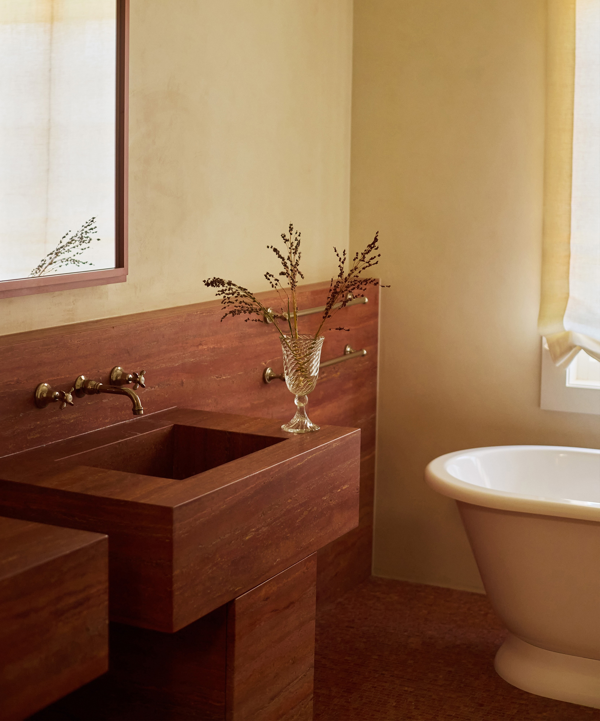 A minimalist bathroom in a Spanish style home features a stone sink, wall-mounted faucet, glass vase with dried flowers, a large mirror, and a white freestanding bathtub near a window with sheer curtains.