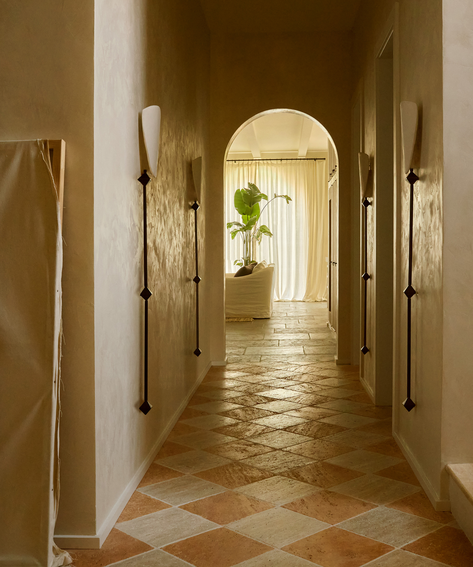 A sunlit hallway with checkered tile flooring leads to a bright living area in this Spanish style home, featuring white curtains, a leafy plant, and a white sofa. Wall sconces line the hallway, and soft light creates a calm atmosphere.