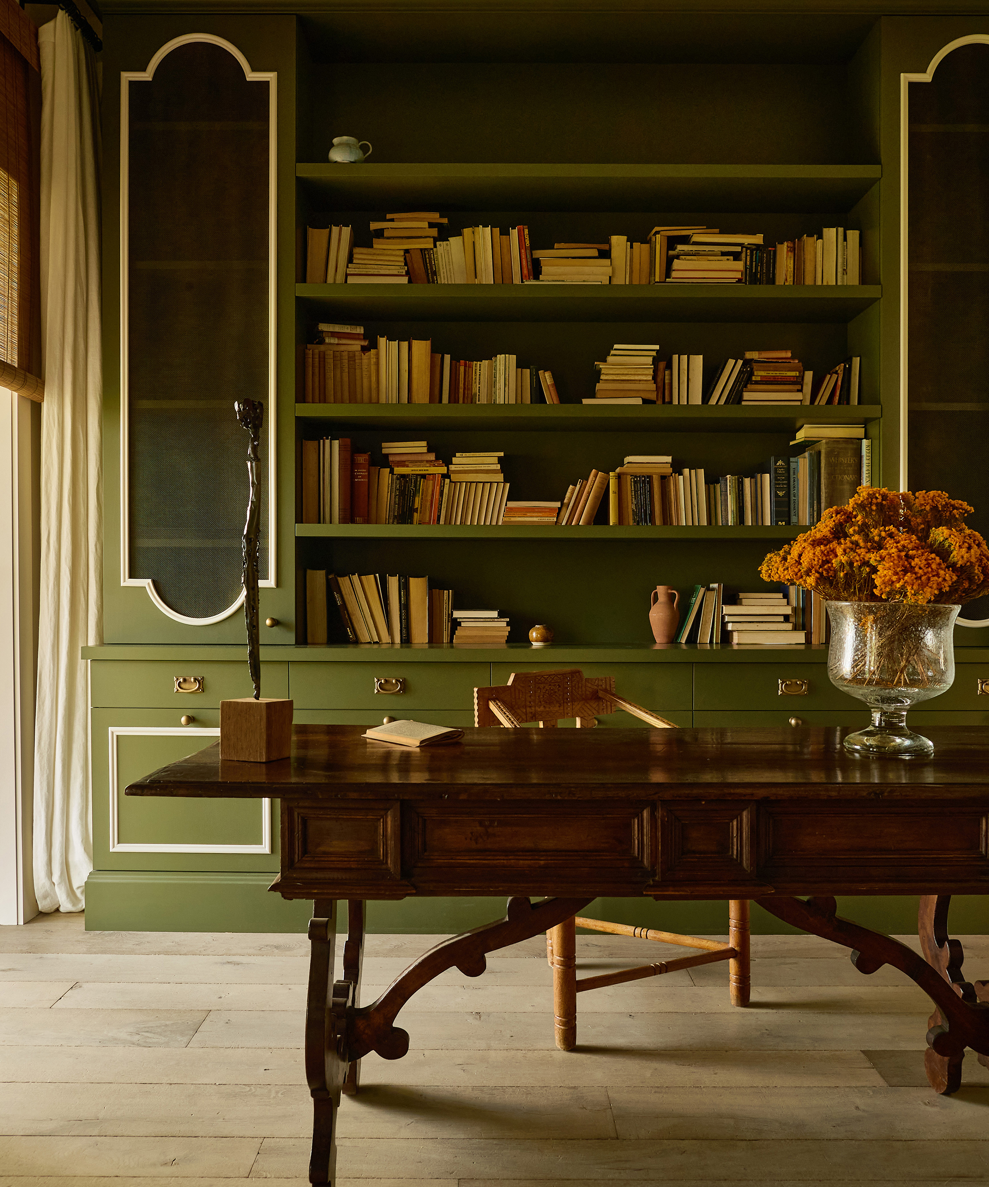A warmly lit study in a Spanish style home features green bookshelves filled with books, a vintage wooden desk, a chair, a flower arrangement in a glass vase, and decorative objects. Light streams in from a window with curtains.