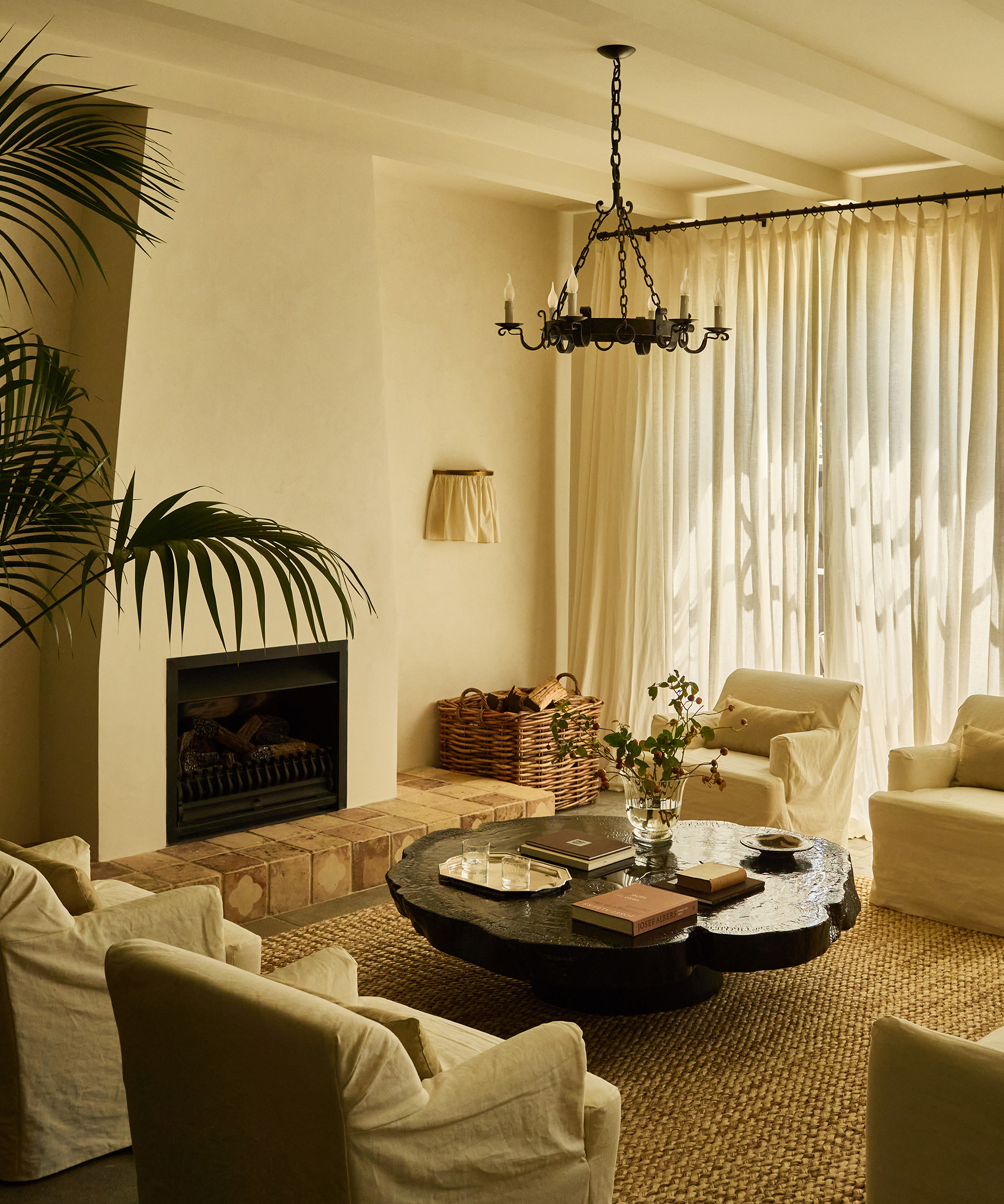 A cozy spanish style home living room with cream-colored armchairs, a round dark coffee table, a built-in fireplace, wicker baskets, sheer curtains, and soft natural light creating a warm, inviting atmosphere.
