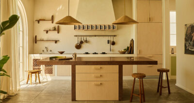 A modern kitchen in a Spanish style home, featuring light wood cabinets, a large island with brown countertops, woven pendant lights, bar stools, open shelves with kitchenware, and natural light streaming in from tall windows.