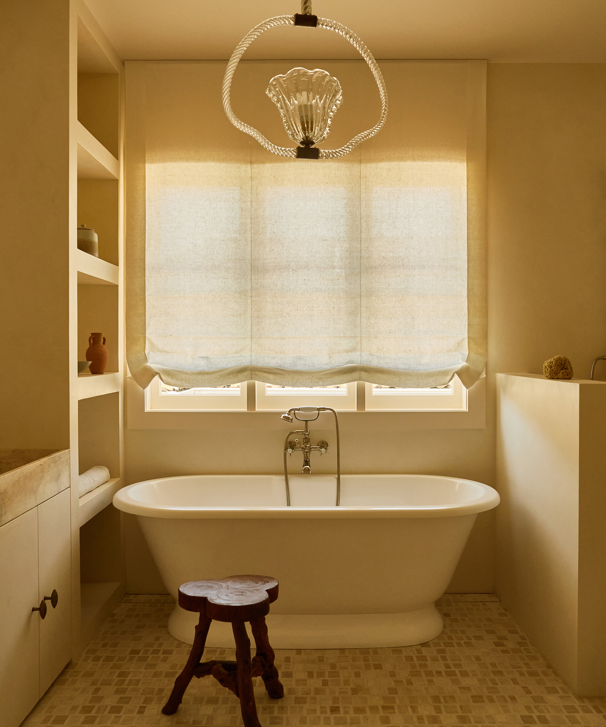 A minimalist bathroom in a Spanish style home features a freestanding white bathtub, a wooden stool, built-in shelves, and a large window with a sheer beige Roman shade. A decorative light fixture hangs from the ceiling above the tub.