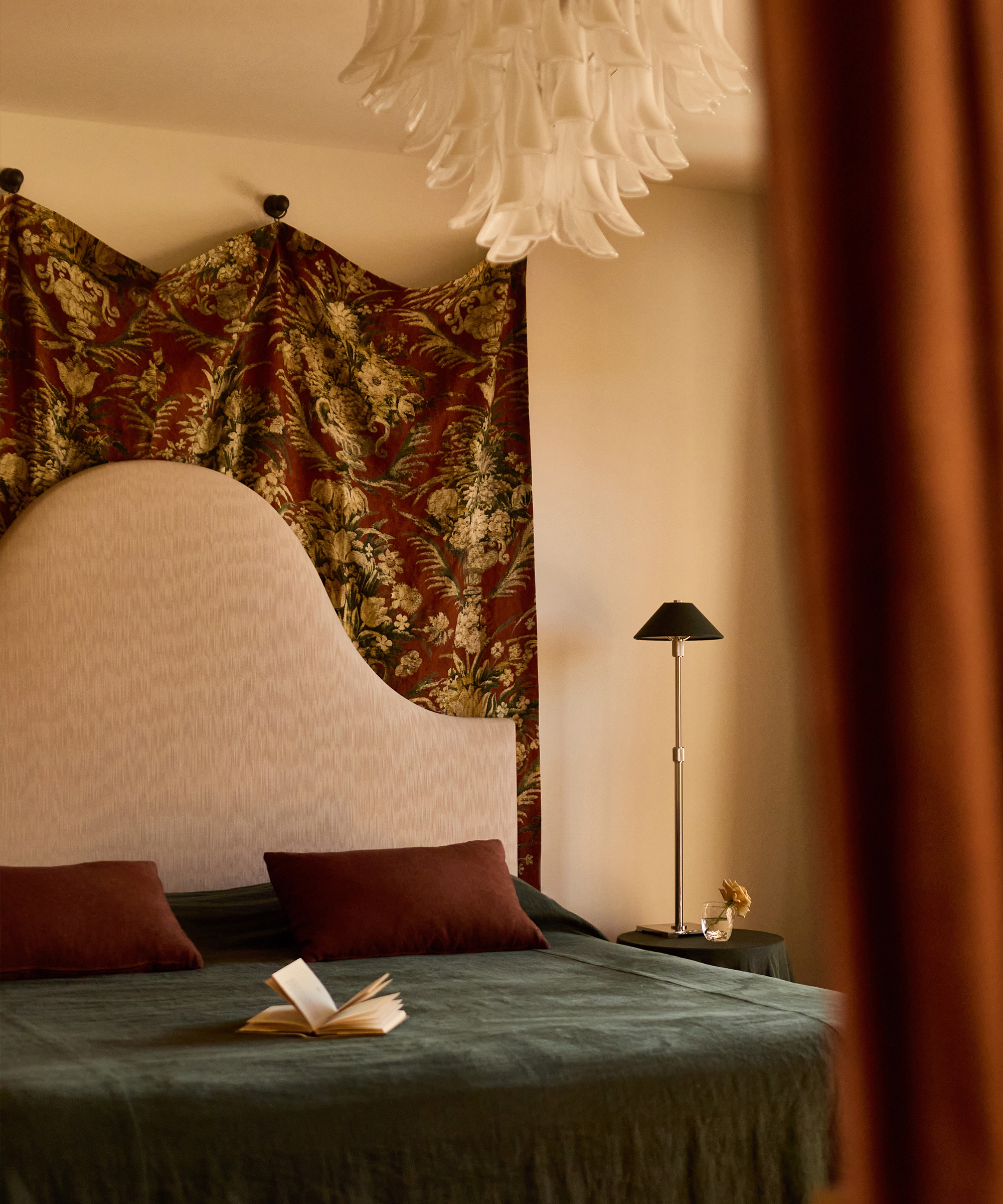 A cozy bedroom in a Spanish style home with a tall, curved headboard, dark green bedding, and two brown pillows. A floral tapestry hangs behind the bed, and an open book rests on the bed beside a modern lamp on a small bedside table.